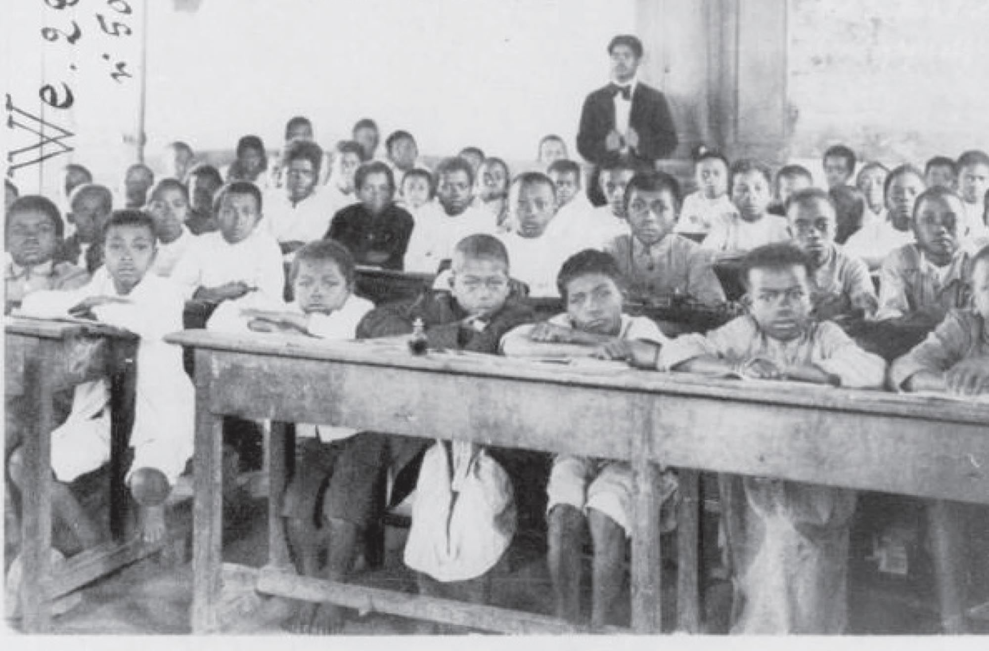 Black and white photograph of a classroom in Madagascar. The children stare straight ahead at the camera and the teacher stands in the background.