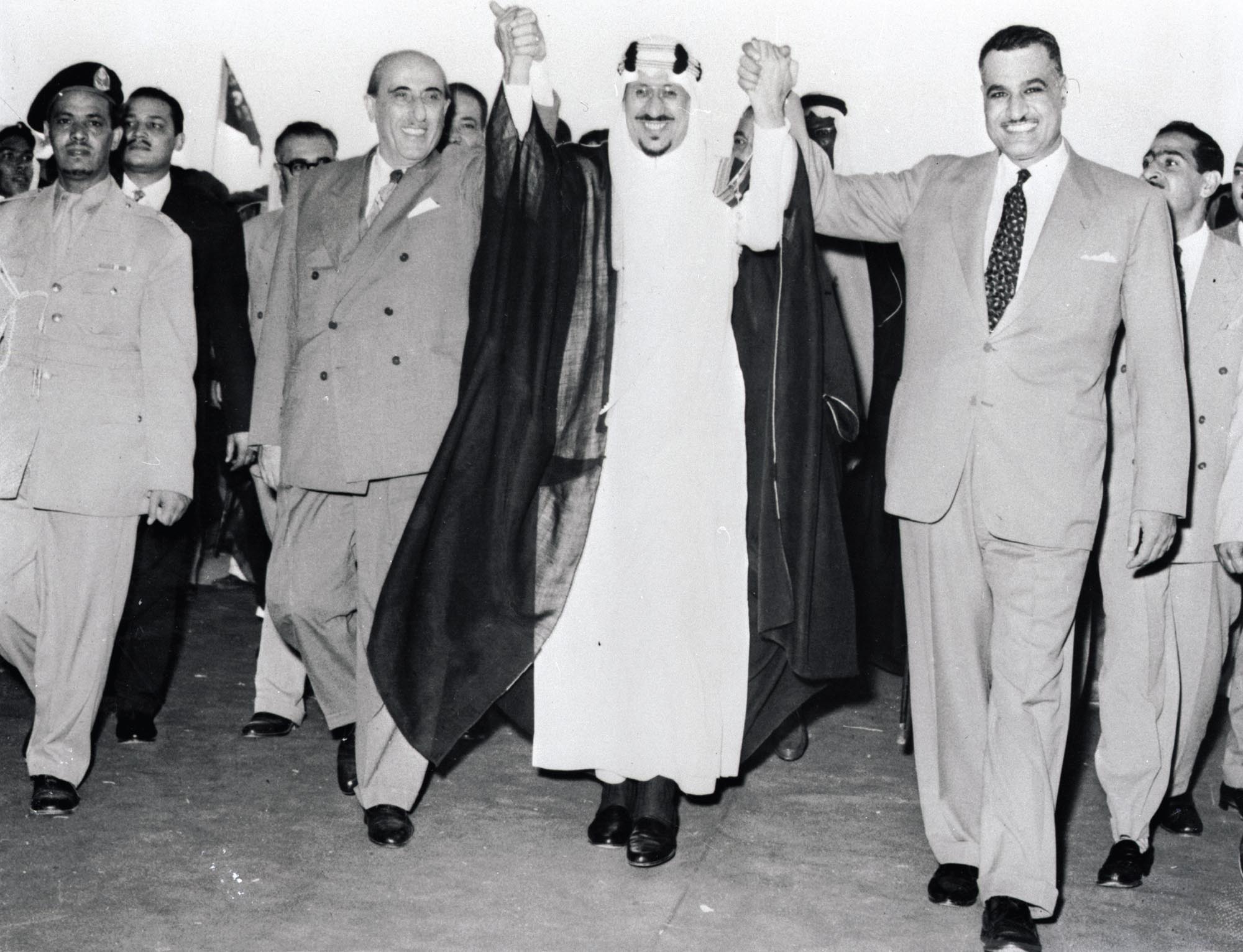 Three Middle Eastern leaders dressed in suits and traditional garments are walking with raised hands and smiling.