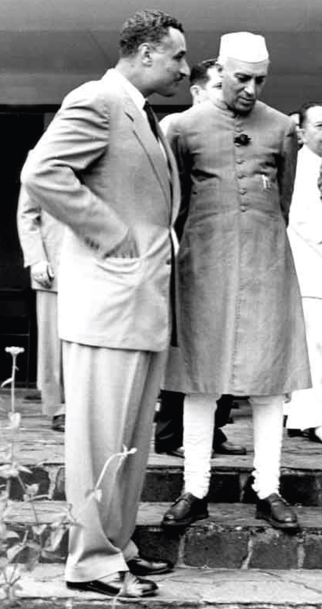 Two men standing together in conversation, the man in a suit has hands in his pockets.