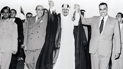 Three Middle Eastern leaders dressed in suits and traditional garments are walking with raised hands and smiling.