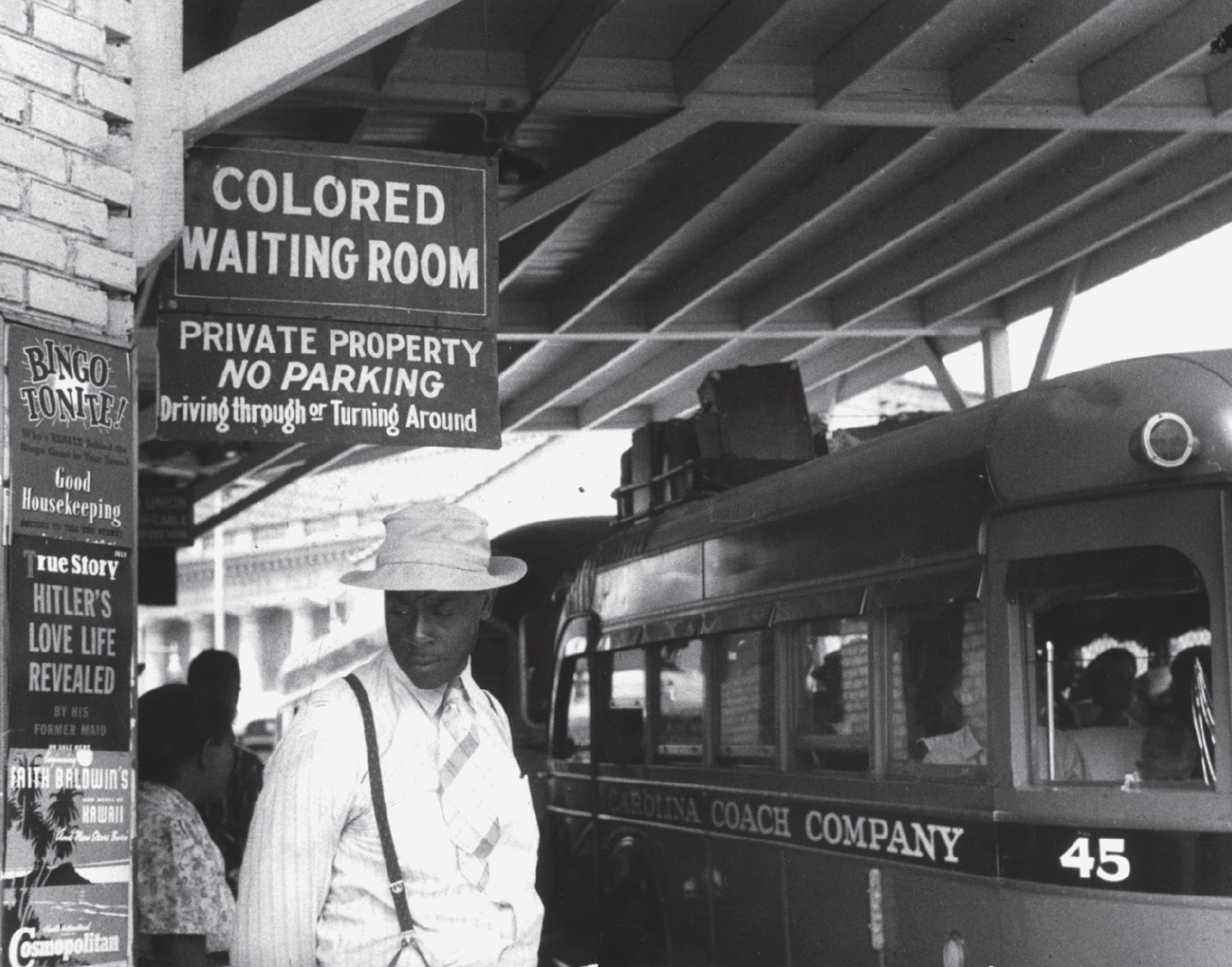 Photo of a Black man at a train station waiting under a sign that says “Colored Waiting Room”.