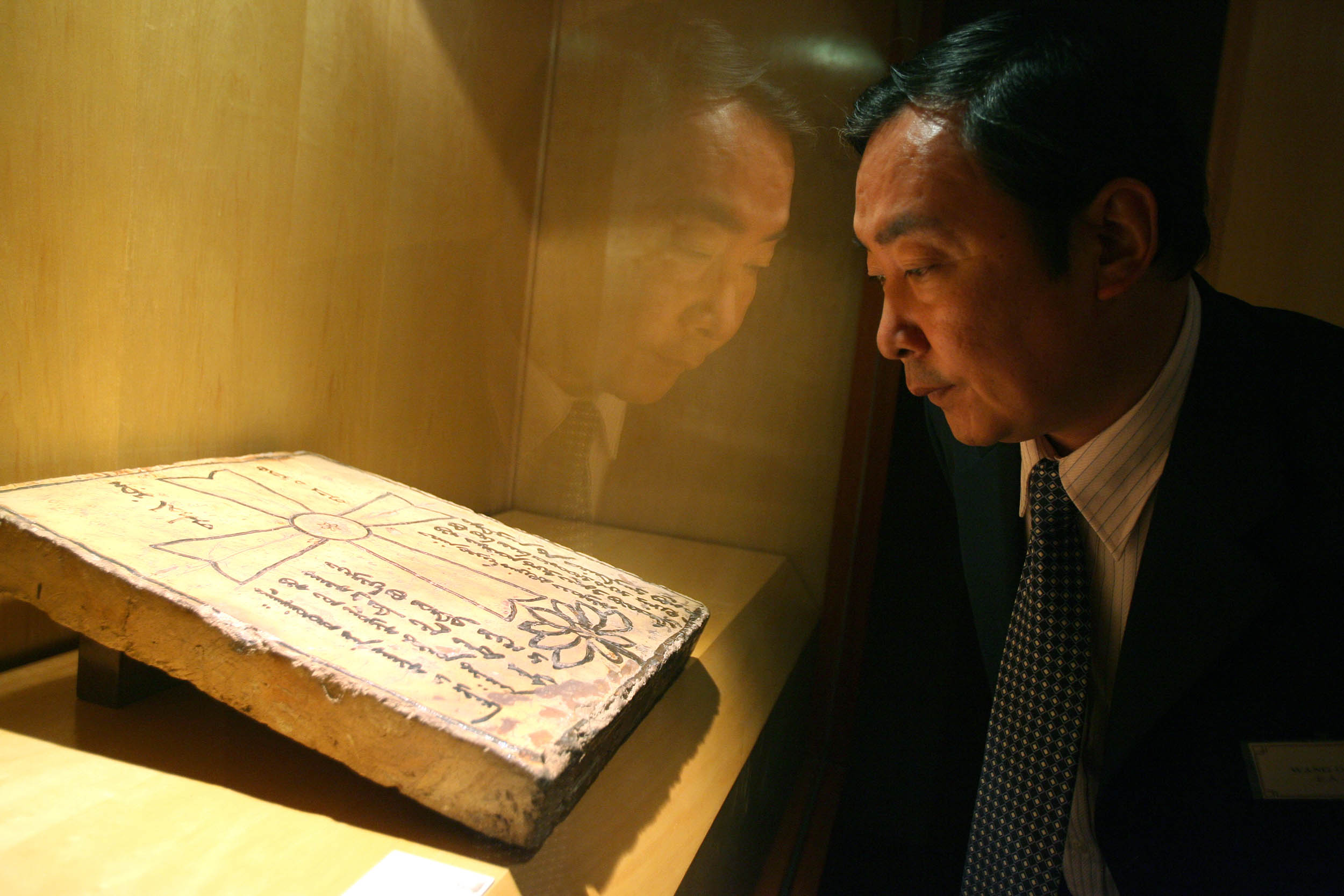 A man in a suit closely examining an inscribed stone tablet displayed in a glass museum case. 