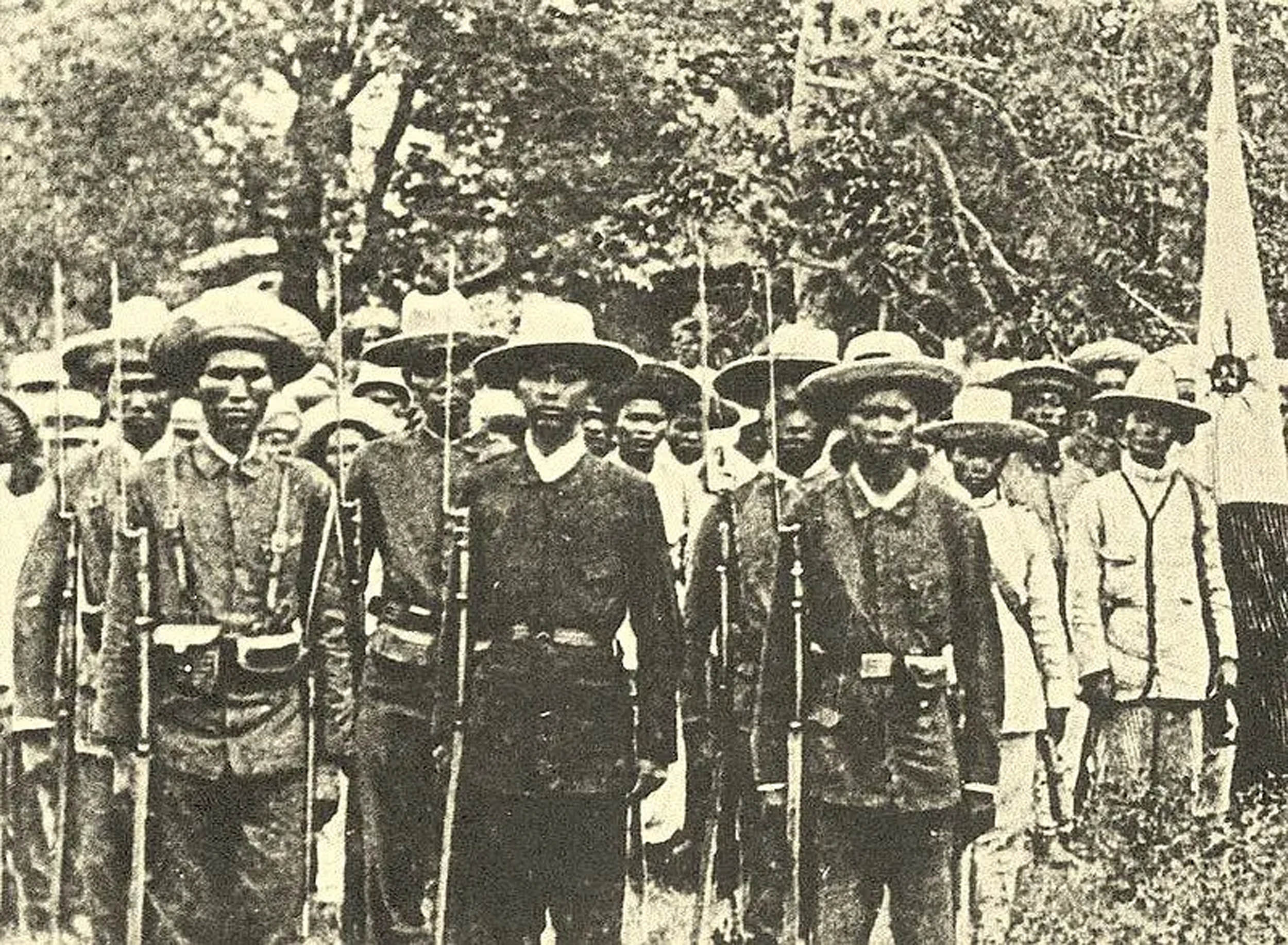 Sepia-toned photograph of a group of uniformed men standing in formation, some holding rifles with bayonets. Most wear wide-brimmed hats and are flanked by others in light-colored clothing, with trees in the background.