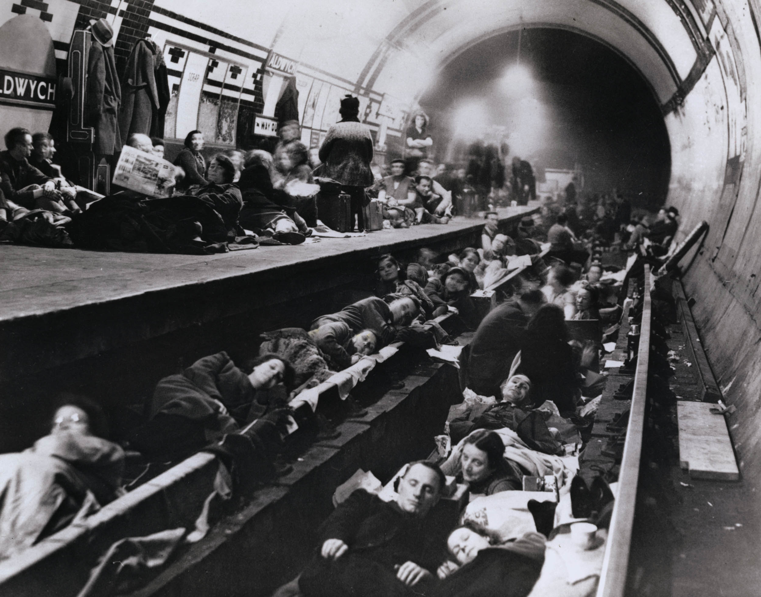 A black and white photo of civilians packed into a London Underground station, lying on the tracks and platform. Blankets and coats cover the sleepers, with people sitting, reading, or watching nearby. Overhead lights cast a soft glow through the tunnel, which fades into the distance. Signs on the tiled wall identify the station as Aldwych.