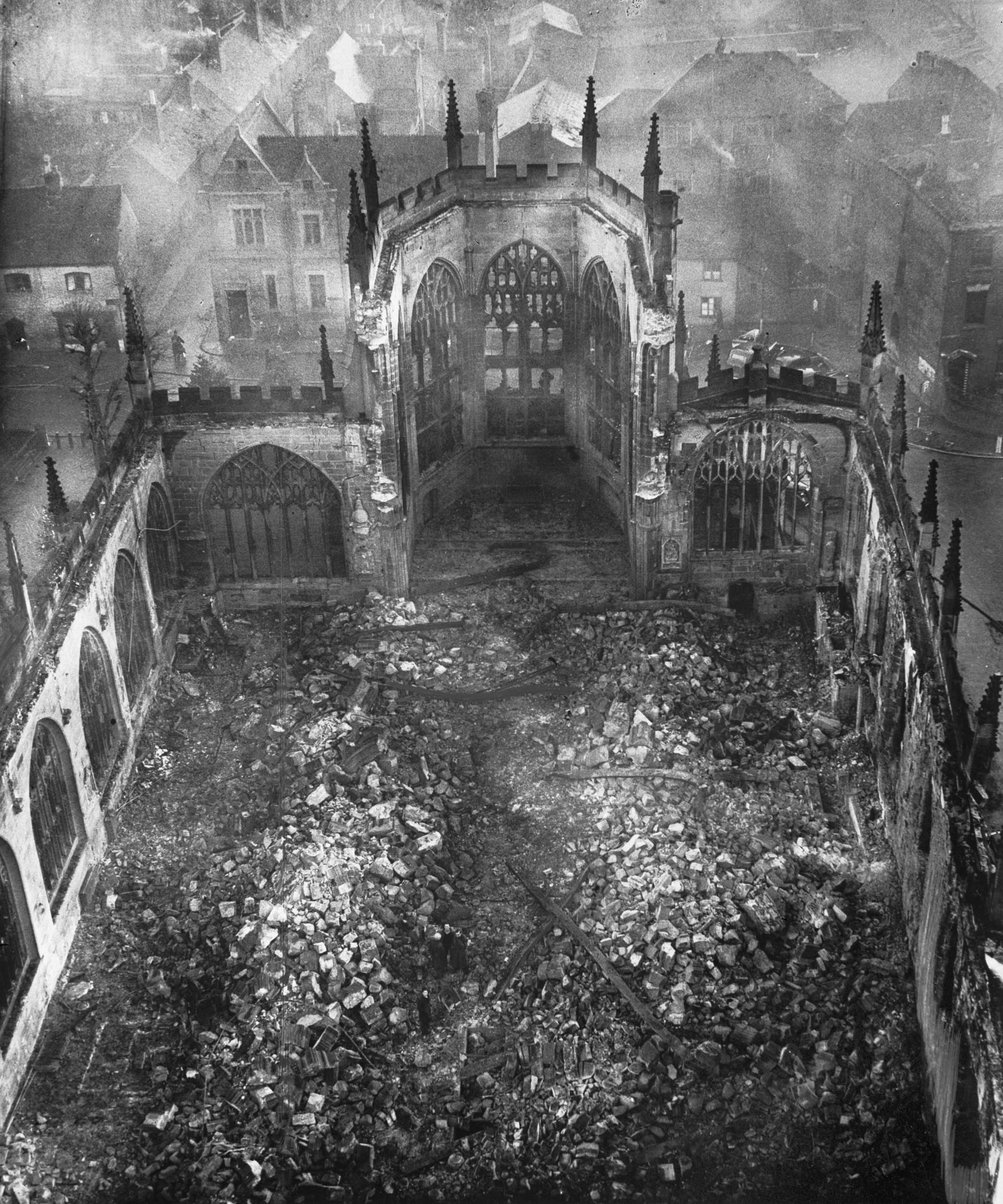 A black and white image of a bombed-out cathedral, with its roof completely destroyed and the interior filled with rubble. Gothic arches and tall windows remain standing, while smoke or haze surrounds the background buildings.