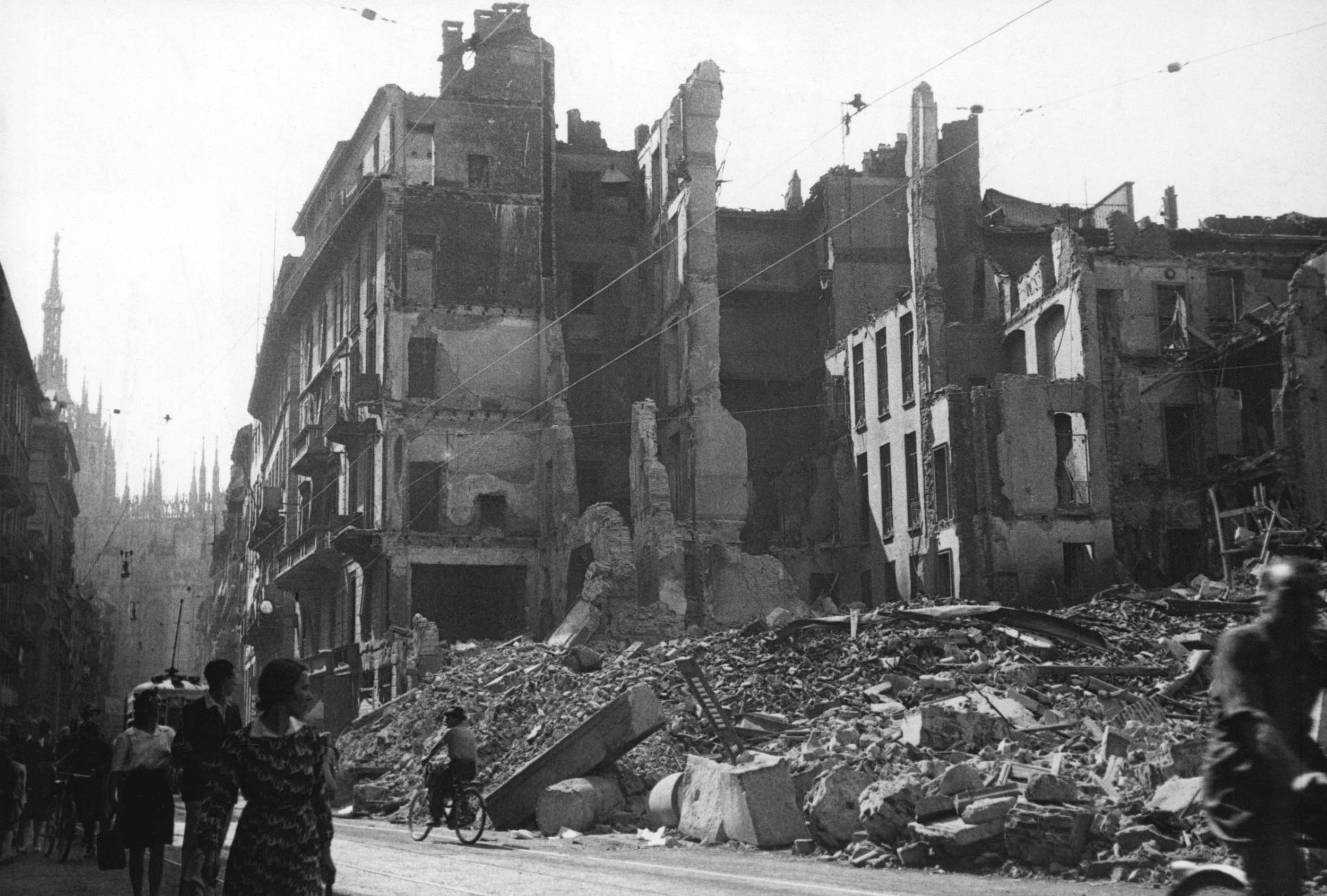 A black and white photo of a heavily bombed urban street. Crumbling buildings line the road, with facades destroyed and interior walls exposed. Large piles of rubble spill into the street, while civilians walk and bike past the ruins.