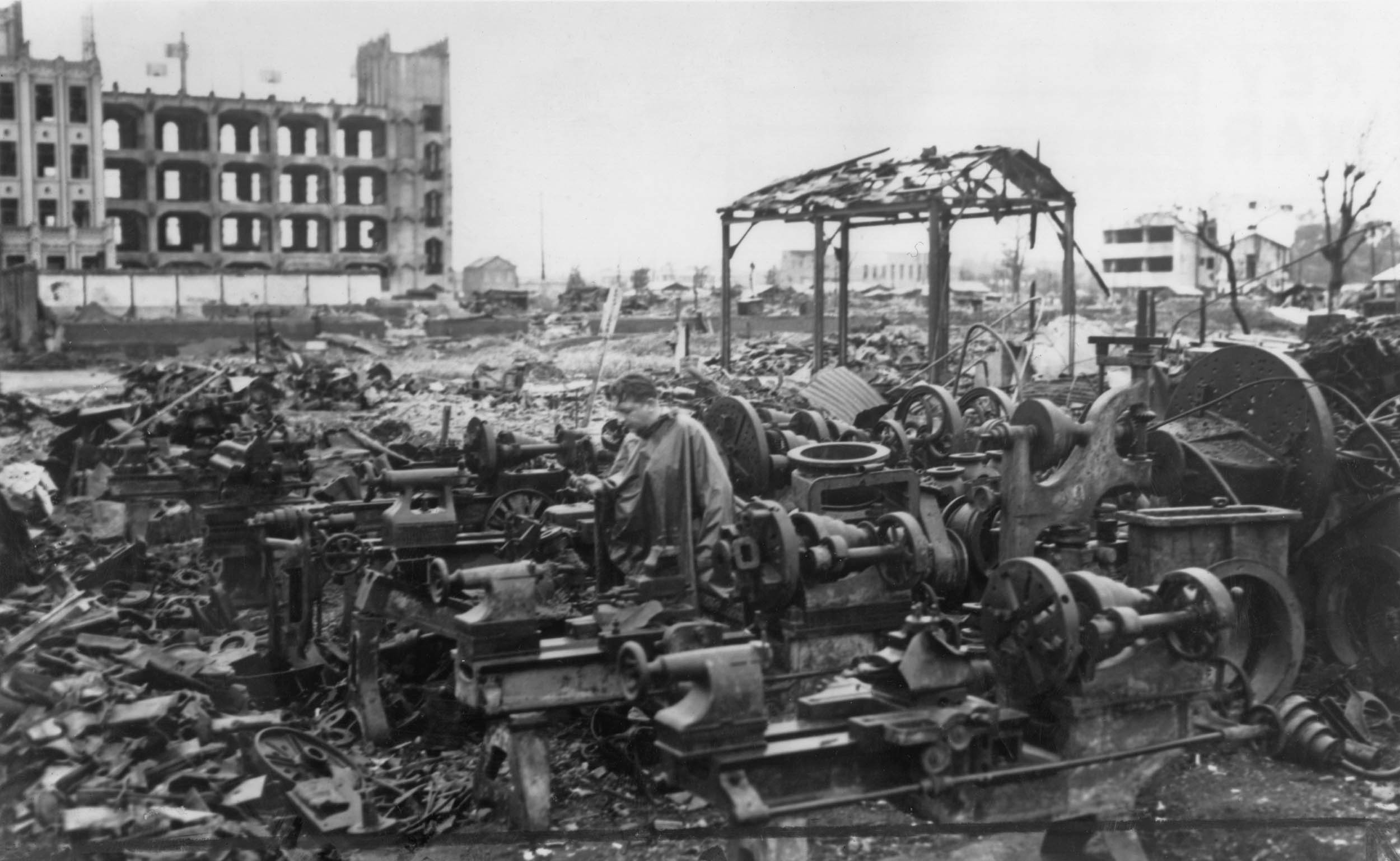 A black and white photo of a bombed-out industrial area. Damaged machinery sits exposed amid debris and twisted metal. A man inspects the ruins, surrounded by the remains of factory equipment. In the background, skeletal building frames and ruined structures highlight the widespread destruction.