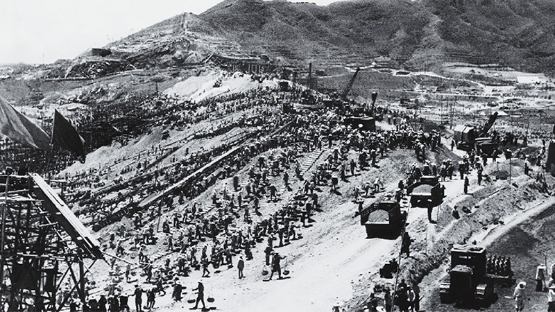 A black-and-white photograph of a large construction site on a hillside, with crowds of workers spread across the slopes and trucks and machinery in the foreground.
