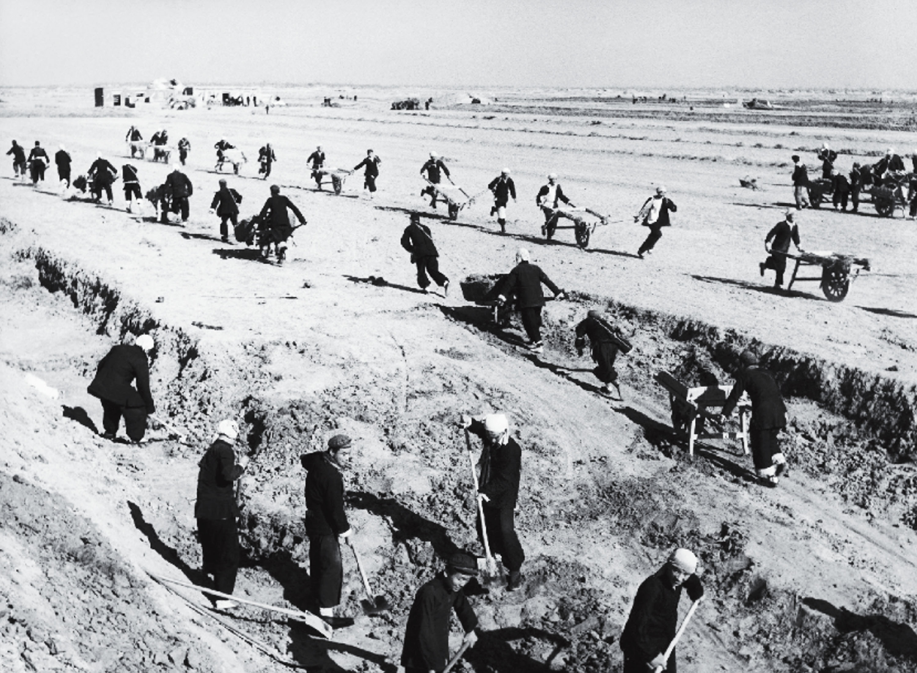 A black-and-white photograph of groups of workers digging trenches in the foreground while others push wheelbarrows across open, flat land in the background.