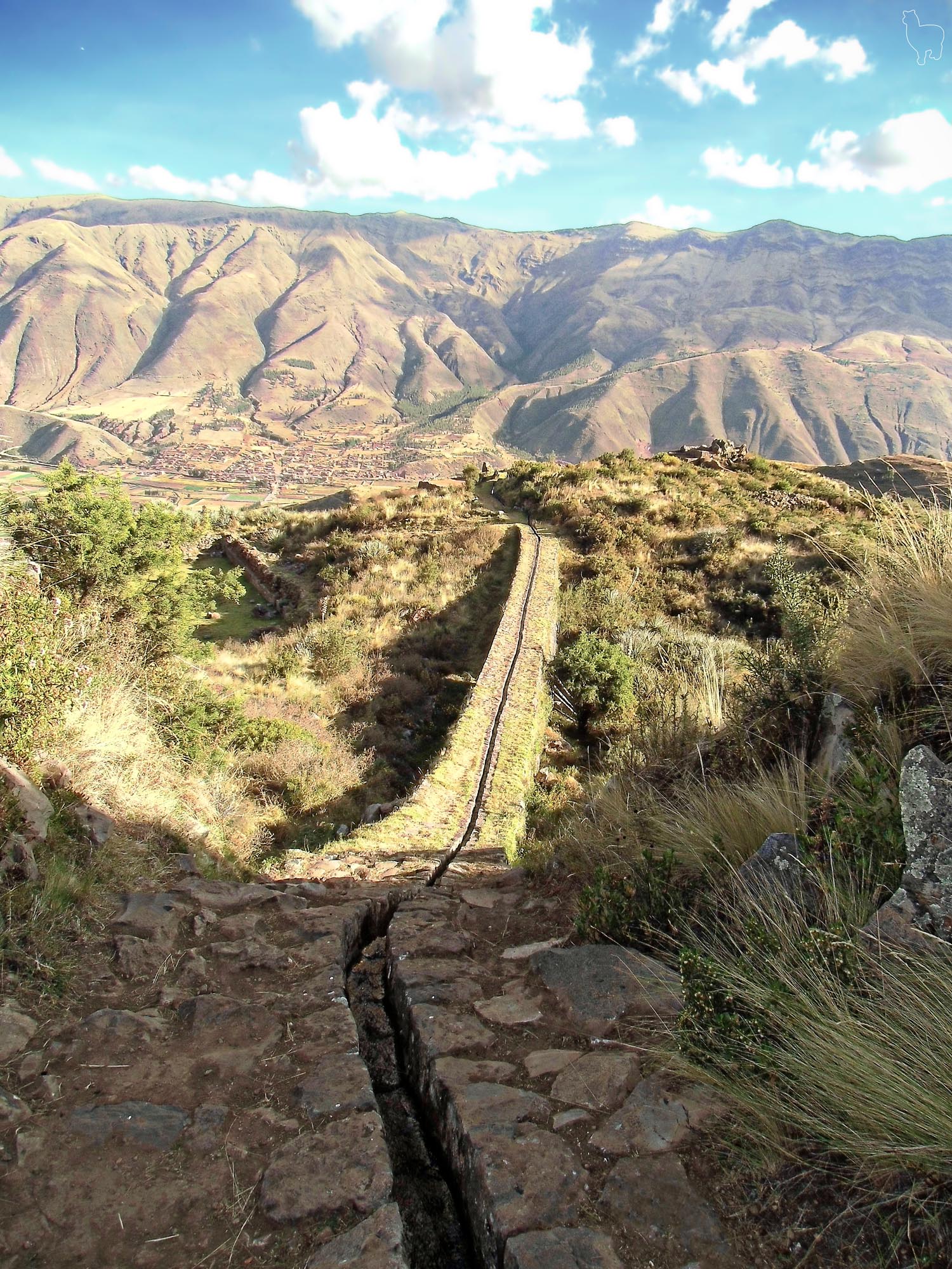 An Incan aqueduct near Cusco, Peru, that winds down a grassy hillside with a valley and mountains in the background under a blue sky with scattered clouds. The stone channel of the aqueduct is visible in the foreground.