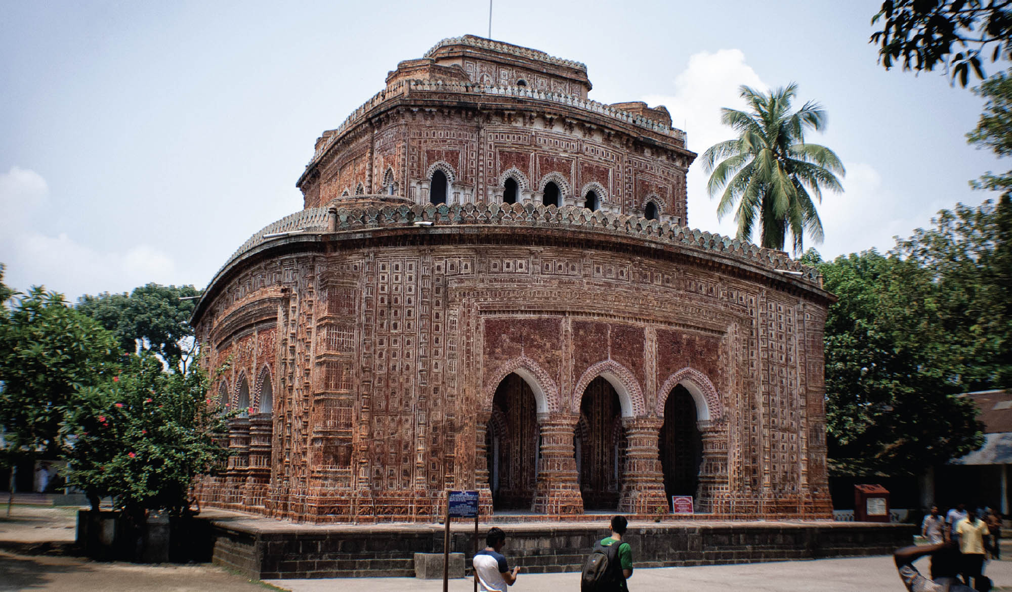 Picture of the Kanatajir Temple in Birgoni, Dinajpur, Bangladesh. A two-story brown stone building with arches entrances and windows in front of a row of trees.