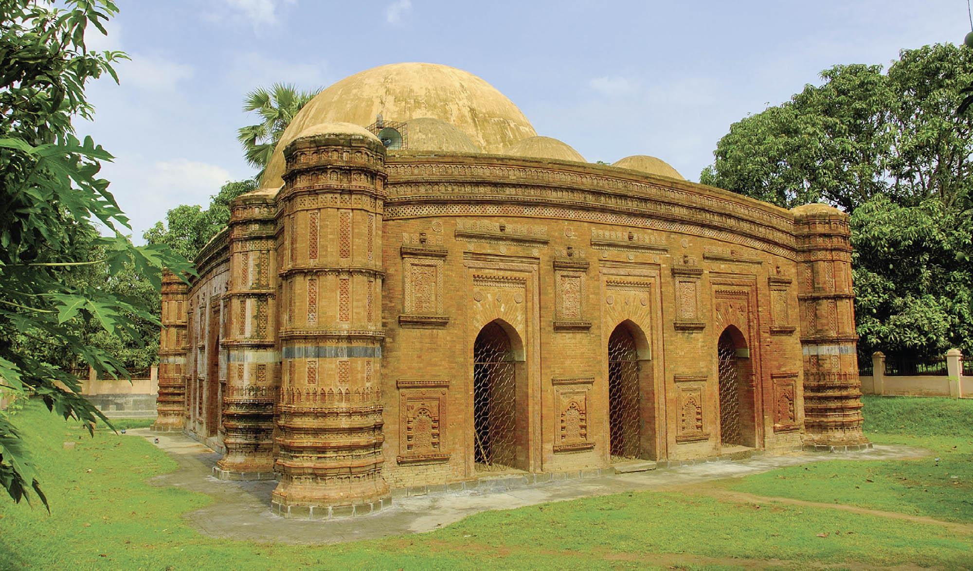 Picture of Khania Dighi Modsque. A one-story mosque with a domed roof and arched entrances in front of a row of trees.