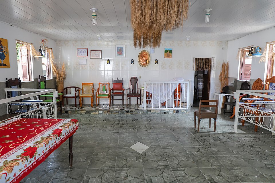 Interior view of a spacious room with a high white ceiling and grey tiled floor, featuring a variety of wooden chairs and benches arranged around a central open space, a red floral tablecloth on a table in the foreground, and a large, textured light fixture made of natural fibers hanging from the ceiling.