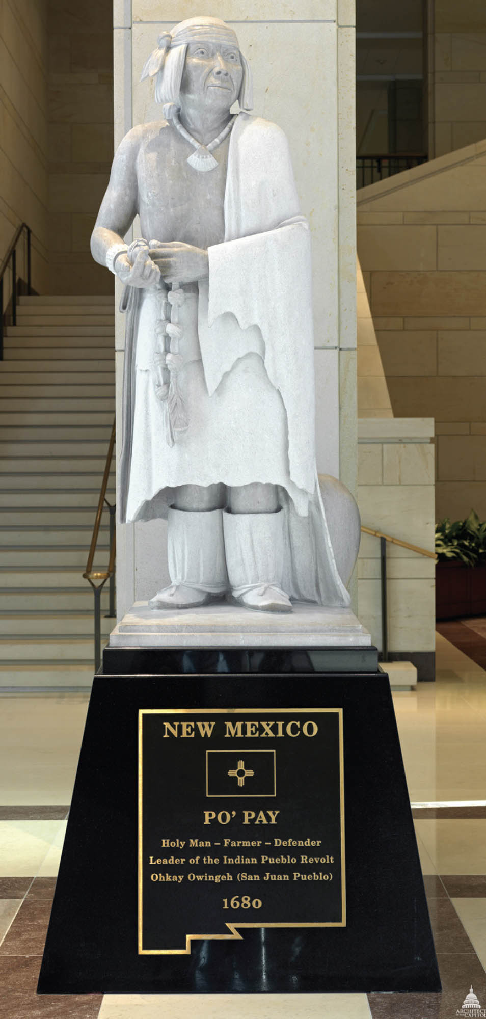 A gray statue of the Popé. The man is wearing boots, a skirt, a necklace, and a wrap, and is holding a string with beads.