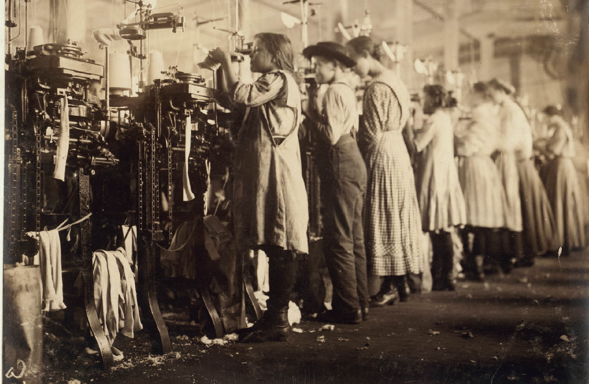 Picture of a row of young people standing up to work on knitting machines in a factory.