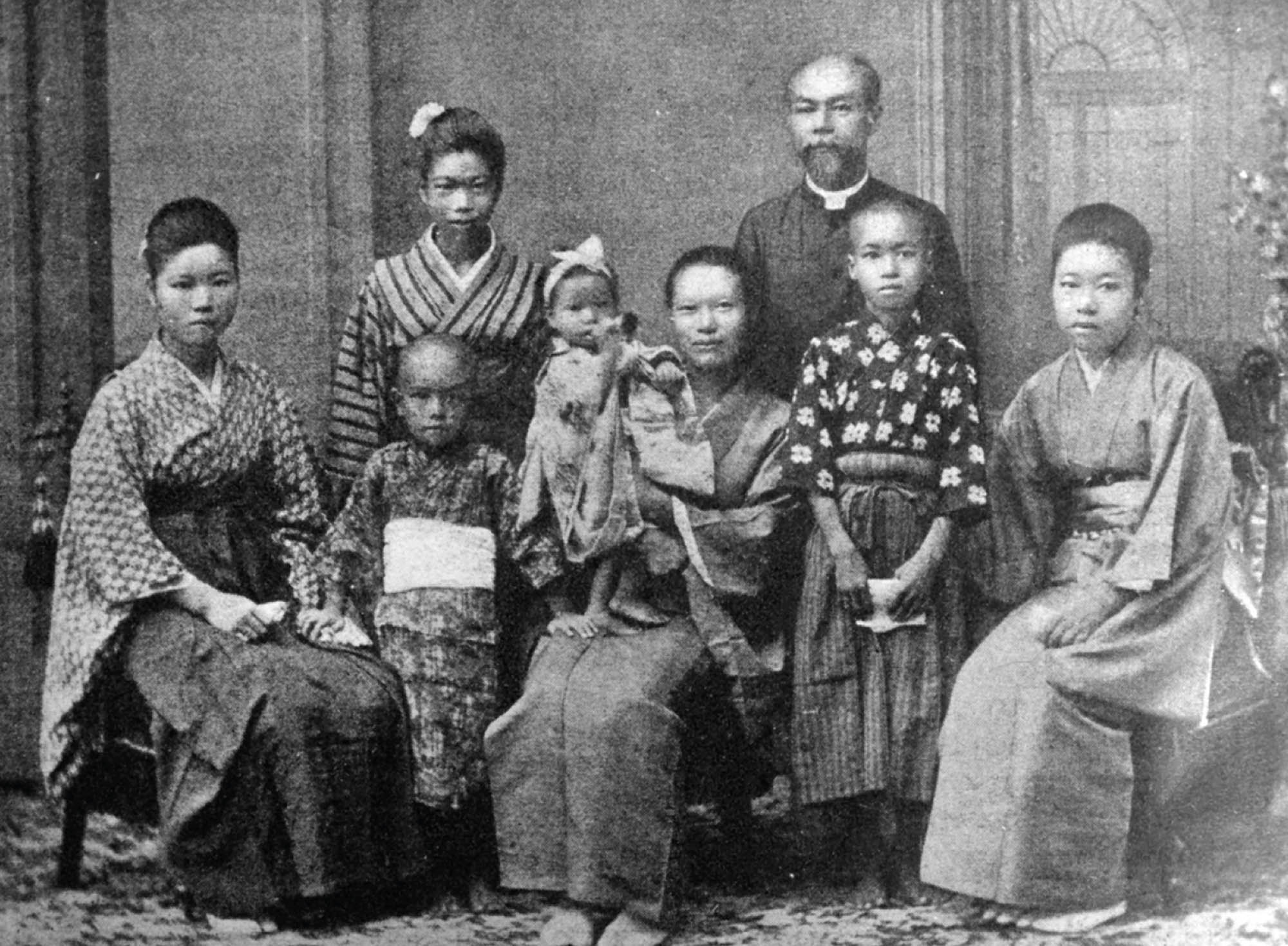 Picture of a family. A older man and a young woman stand behind two standing children. On the right, left, and center sit three women, the middle one with a young child in her arms.