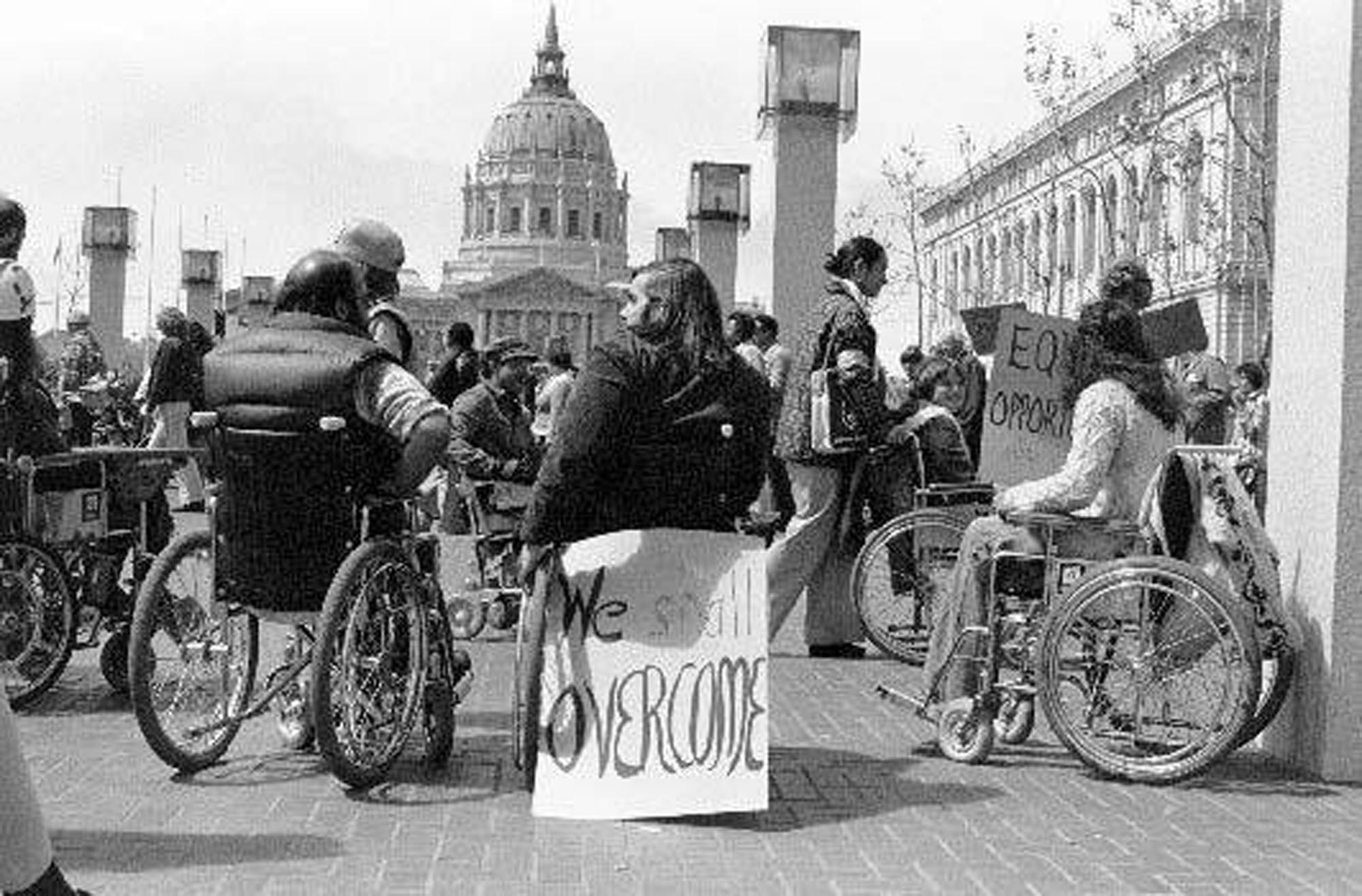 This black and white photograph shows disability activists, many in wheelchairs, gathered outside a building, with protest signs visible, including one proclaiming "We shall OVERCOME". In the background, a domed building, is visible, underscoring the political context of the demonstration.
