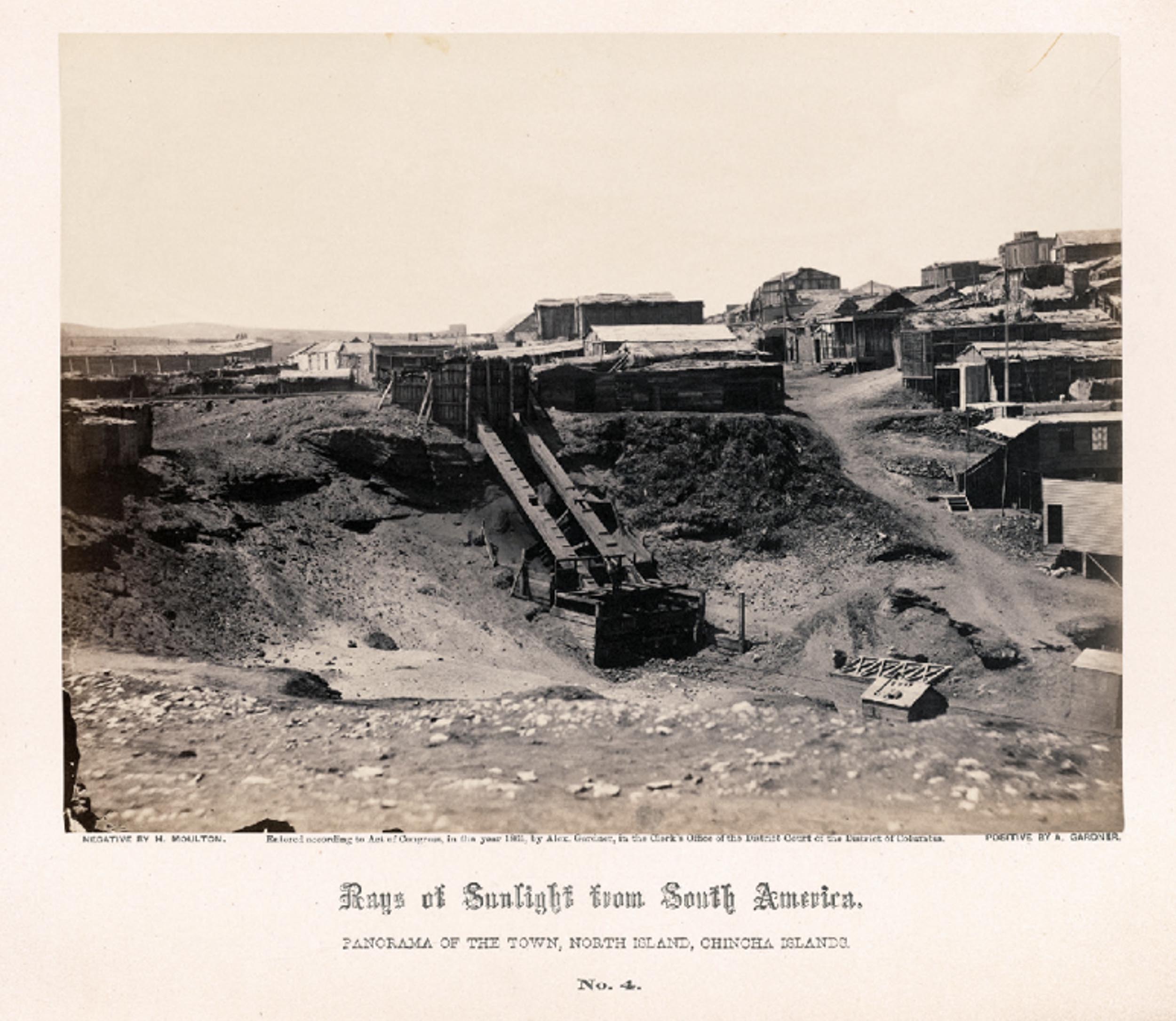 An image from the series, Guano mines in Peru: panoramic view of a the mining town. Rough wooden buildings on a hill, conveyor belts run down to train tracks.