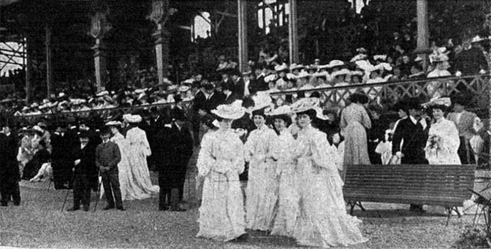 A black and white photograph depicting a large gathering of well-dressed people, primarily women in elaborate hats and long dresses, and men in suits, at what appears to be a formal outdoor. Many people are seated in grandstands in the background, while several individuals are standing and conversing in the foreground.