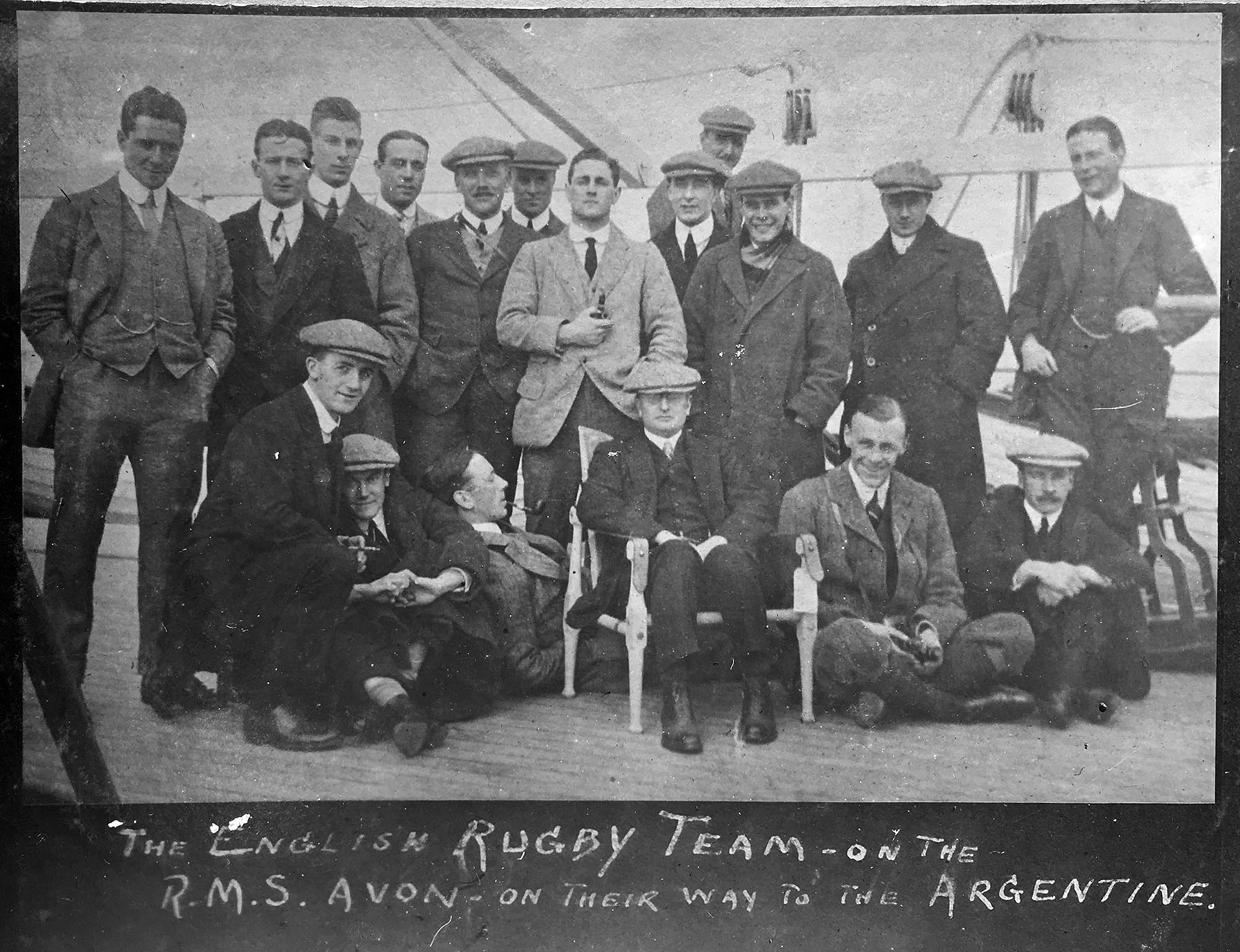 A vintage black and white photograph of the English Rugby Team on the R.M.S. Avon, on their way to Argentina for the 1910 British Lions tour. The team, consisting of about 18 men in suits and caps, pose on the ship's deck, with some standing and others seated or kneeling.