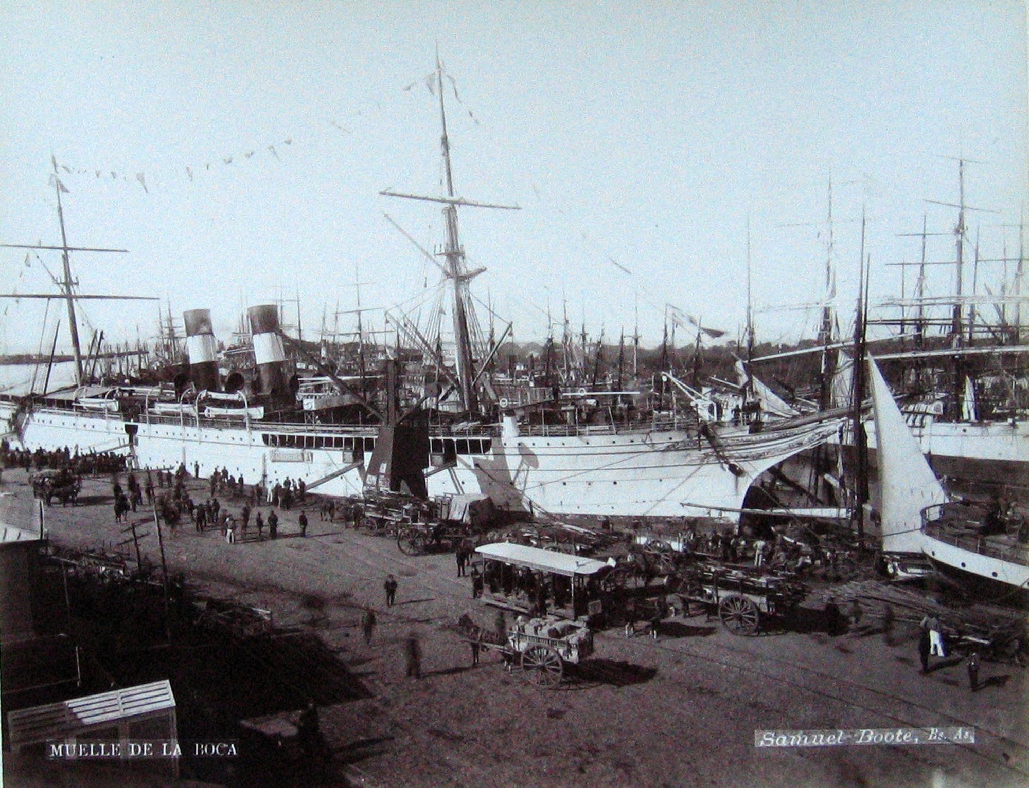 A photograph showing a bustling port scene in Buenos Aires, Argentina, with large ships docked alongside a wharf where people, horse-drawn carts, and a tram are visible, under a bright sky.