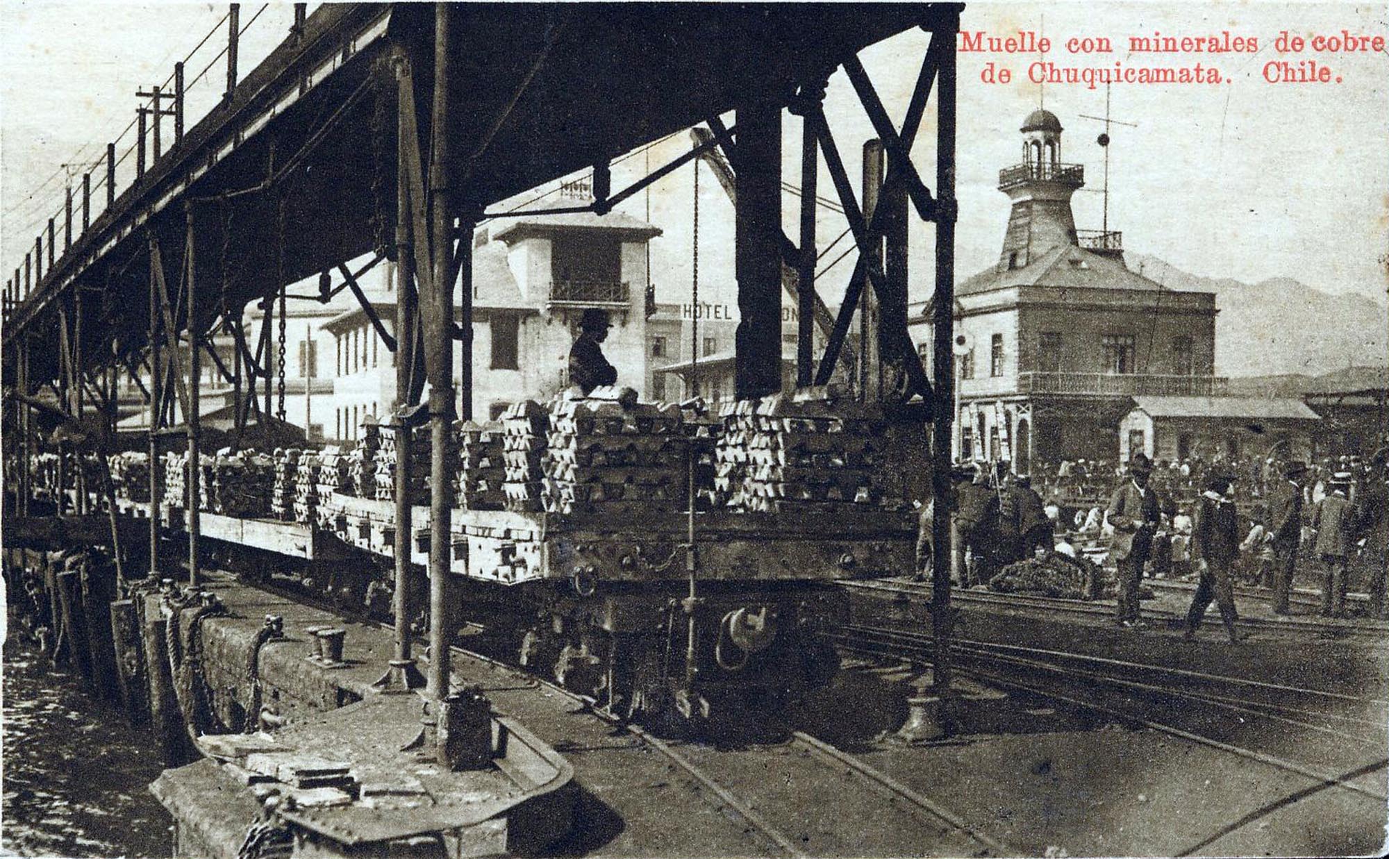 A photograph depicting a pier in Chuquicamata, Chile, where copper minerals are on rail carts. The image shows a long pier structure with a train track running along it; people are visible on the pier, and a building and lighthouse-like structure are visible in the background.
