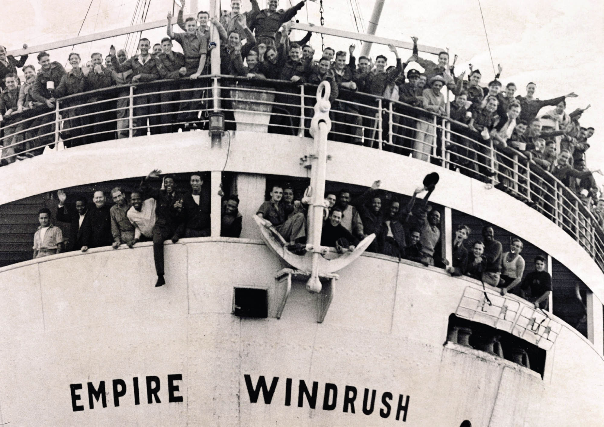 Image of a crowd of men waving from a ship, crowding the railing of the upper deck and around the windows near the anchor. Below them is the name of the ship, Empire Windrush.