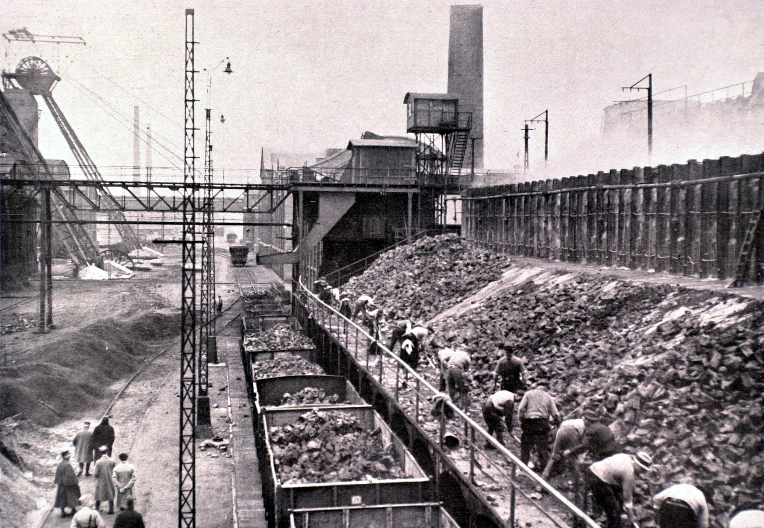 Black and white photo of a large industrial site where workers are loading coal into train cars along a rail line. Smokestacks, machinery, and factory buildings are visible in the background.