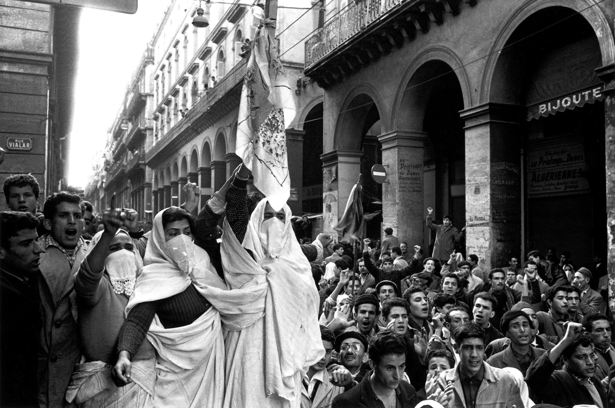 Protestors fill the streets of an Algerian city to protest the colonization and the Cold War. Women wear traditional clothing wave flags in support of liberation.