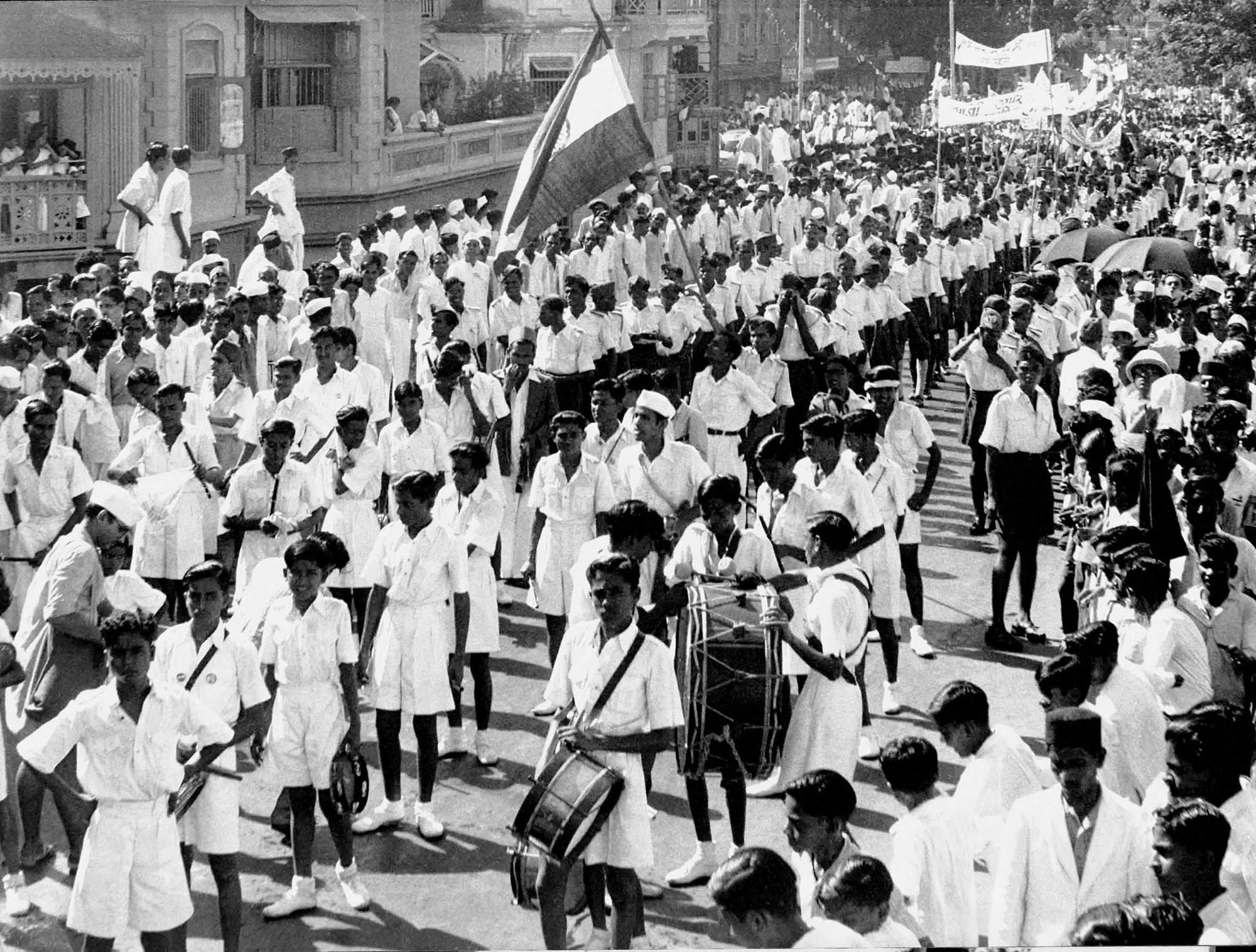 A crowd of people march in the street in long lines. Some in the parade through carry flags and others drums to celebrate the First Independence Day of India.
