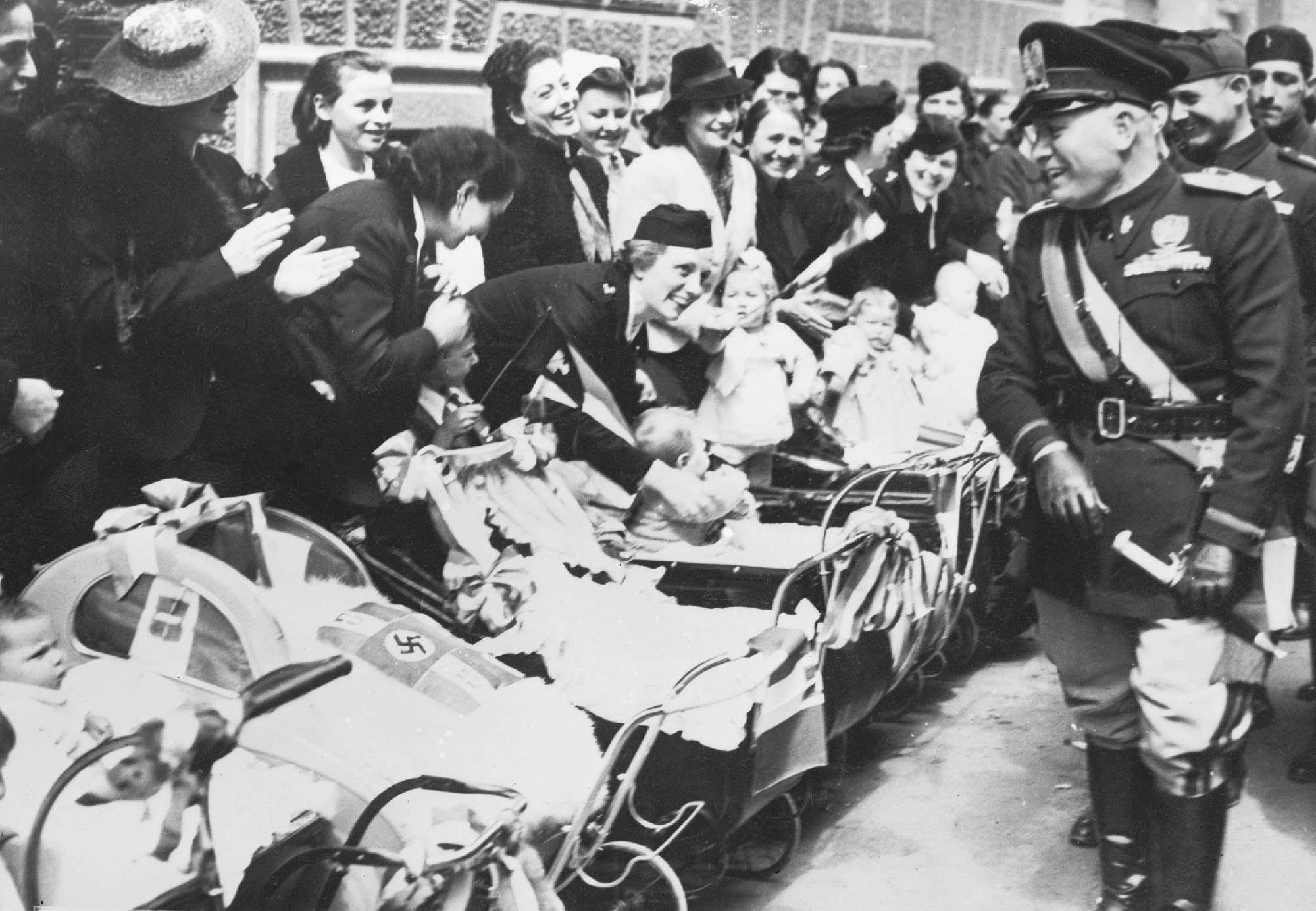 Photograph of several smiling women, pushing prams and/or carrying babies. Next to them are several smiling soldiers. One of the prams has a swastika decal on it.