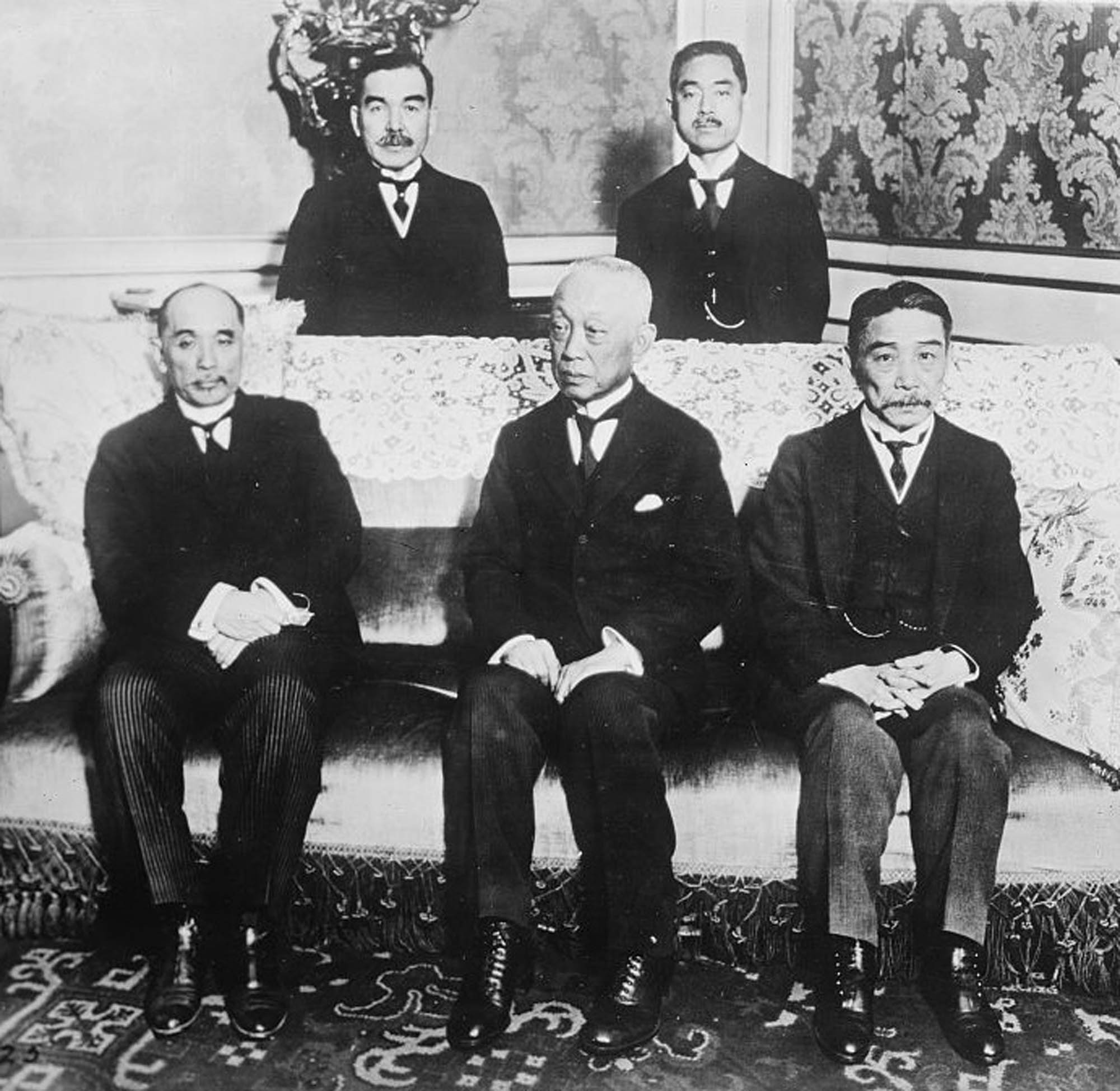 Photograph of five men inside an ornately decorated room. Three are seated on a couch, and two other stand behind them.