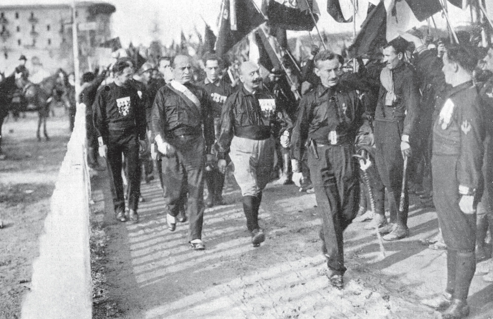 Photograph of a group of men marching down the street. Next to them, a crowd waves flags and salutes.