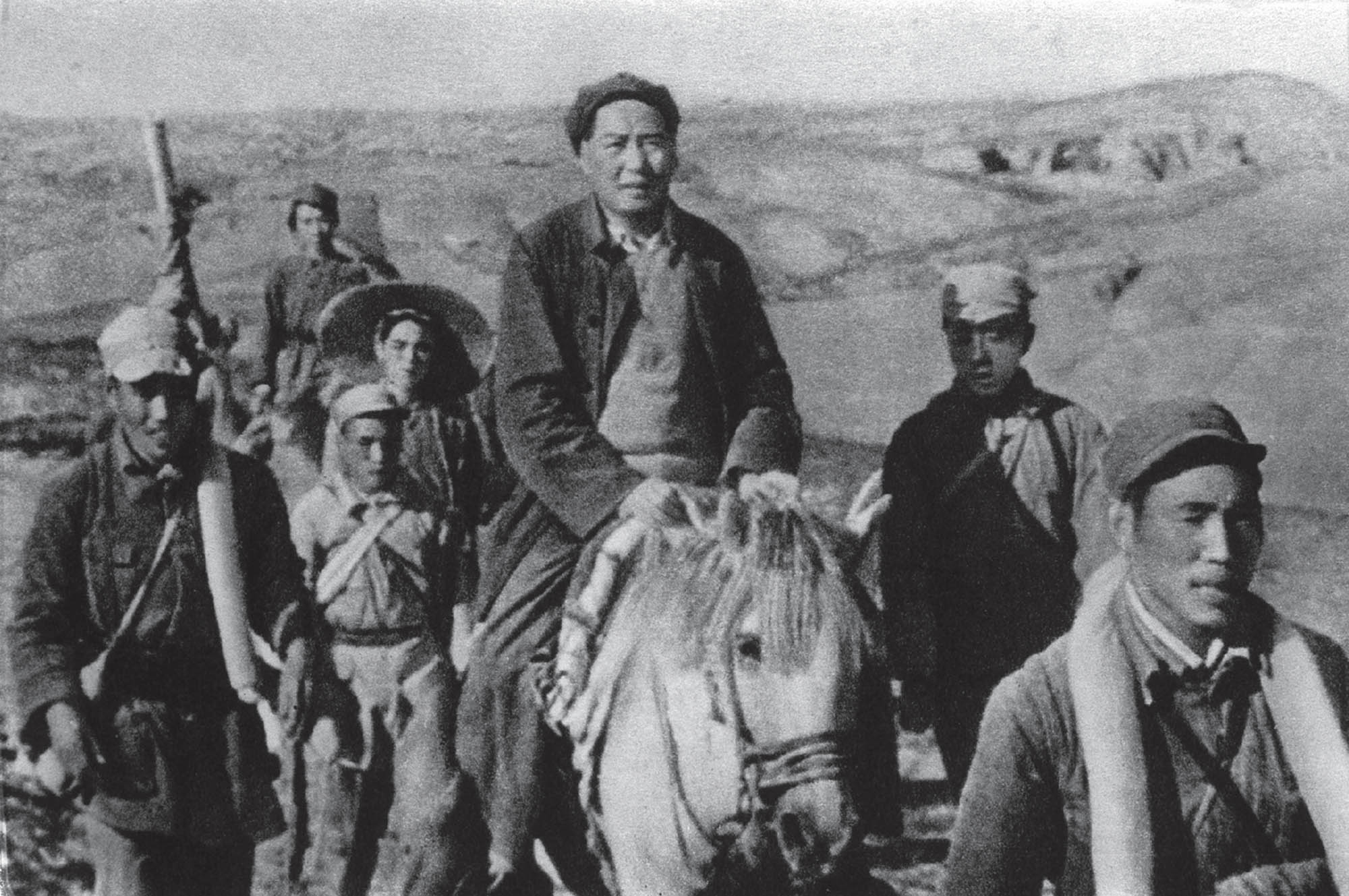 Black and white image of a man on horseback surrounded by other men, all walking through the mountains.