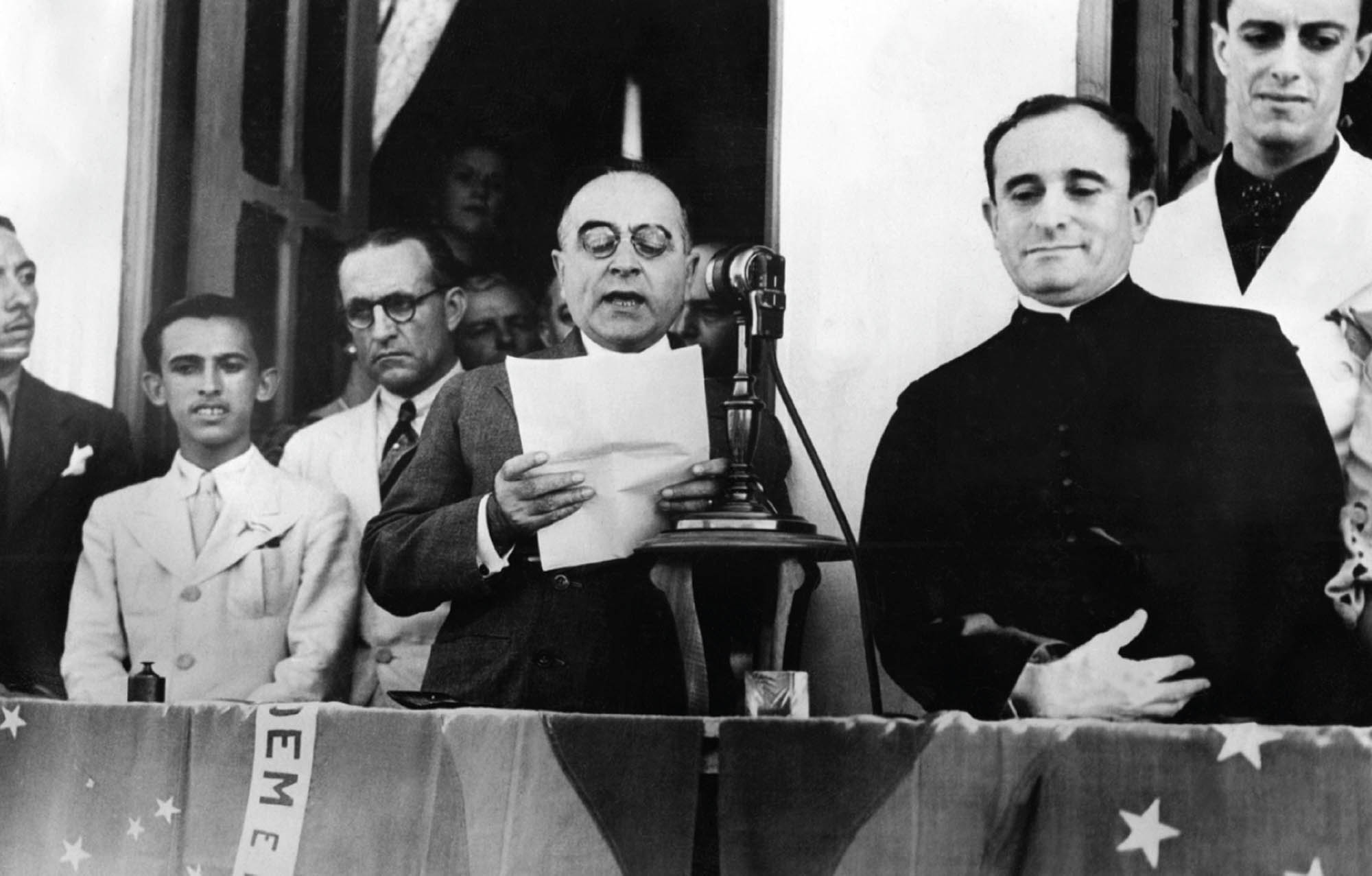 Black and white image of a man giving a speach in front of a microphone, reading from a sheet of paper. Two man stand to the left of him and a small group of people stand behind him to the left.