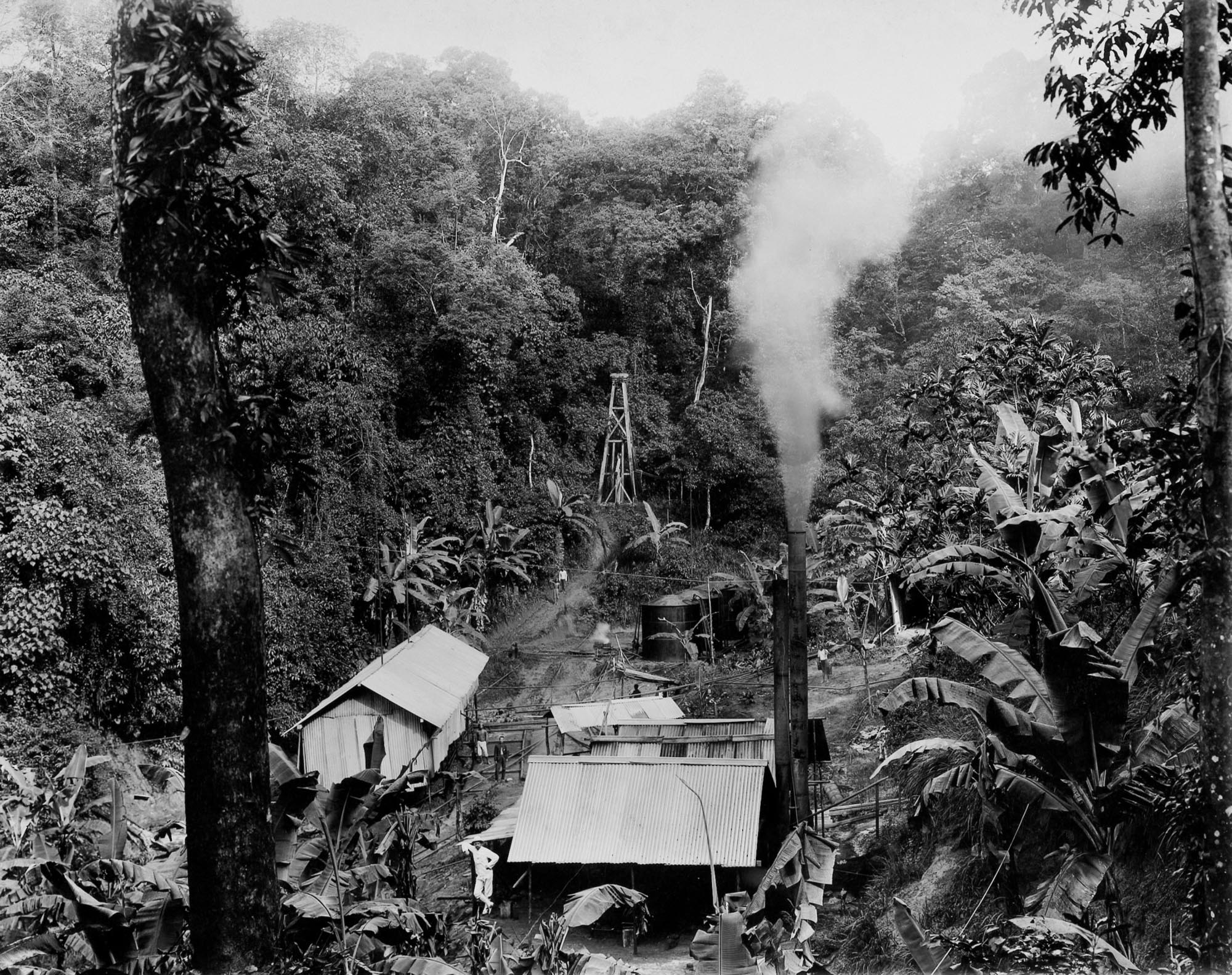 A black and white photo showing an early industrial site nestled within a dense jungle environment. Several simple structures with corrugated roofs are visible, along with a tall smokestack emitting a plume of smoke.
