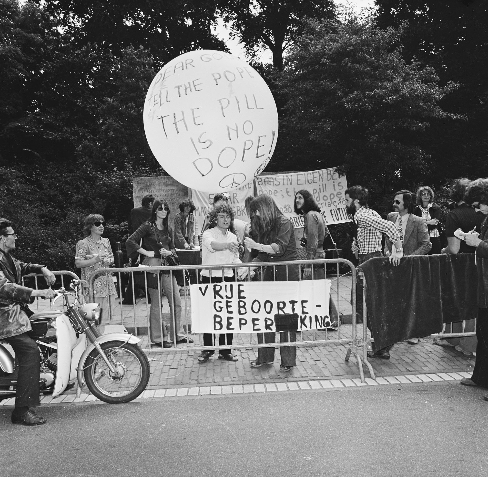 A group of people gathered around a large white balloon and banners. The prominent balloon, held aloft by a pole, displays the text "THE PILL IS NO DOPE!" Another banner held by protesters reads "VRIJE GEBOORTE-BEPERKING". Individuals are visible standing behind metal barriers.