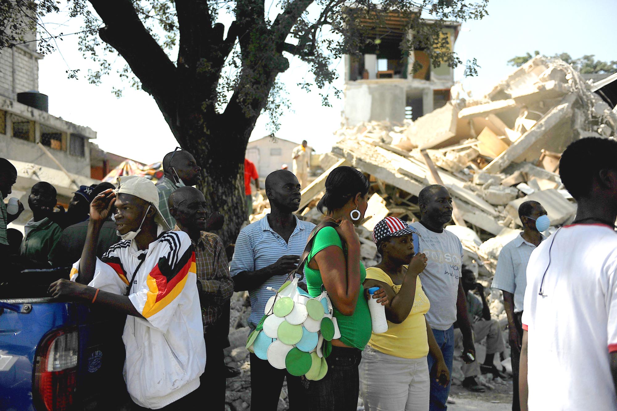 A group of Haitian residents, some wearing masks, stand near a blue pickup truck amidst the rubble and collapsed buildings in the aftermath of an earthquake in Port-au-Prince, Haiti.