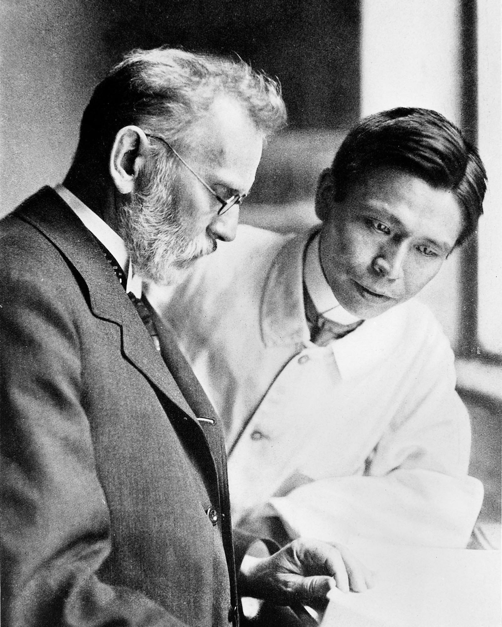 Black and white photograph of an older man with a beard and glasses, looking down at papers held by a younger man, who is wearing a lab coat and also looking at the papers.