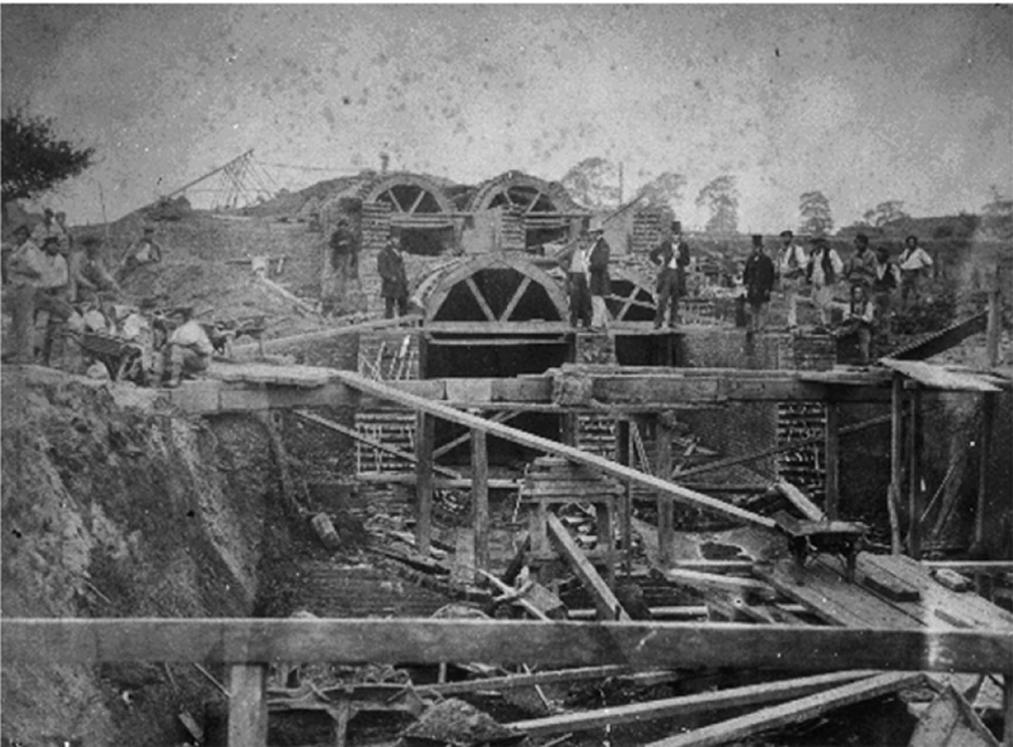 A black and white historical photograph showing multiple workmen on a large construction site with excavated earth and scaffolding. In the foreground, wooden structures and supports are visible, extending into a trench. In the midground and background, several large, circular brick structures are being built.