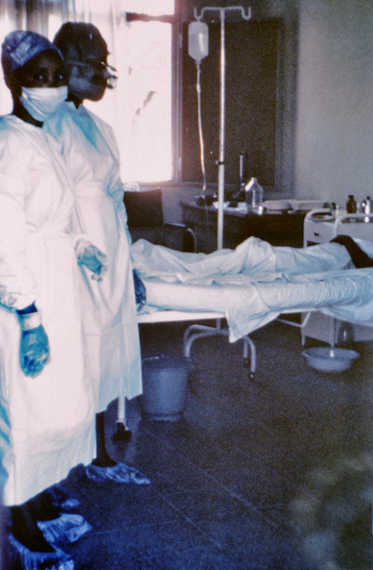 Two nurses in protective gear, including masks, gowns, and gloves, standing in a hospital room next to a bed with a patient being treated for Ebola.