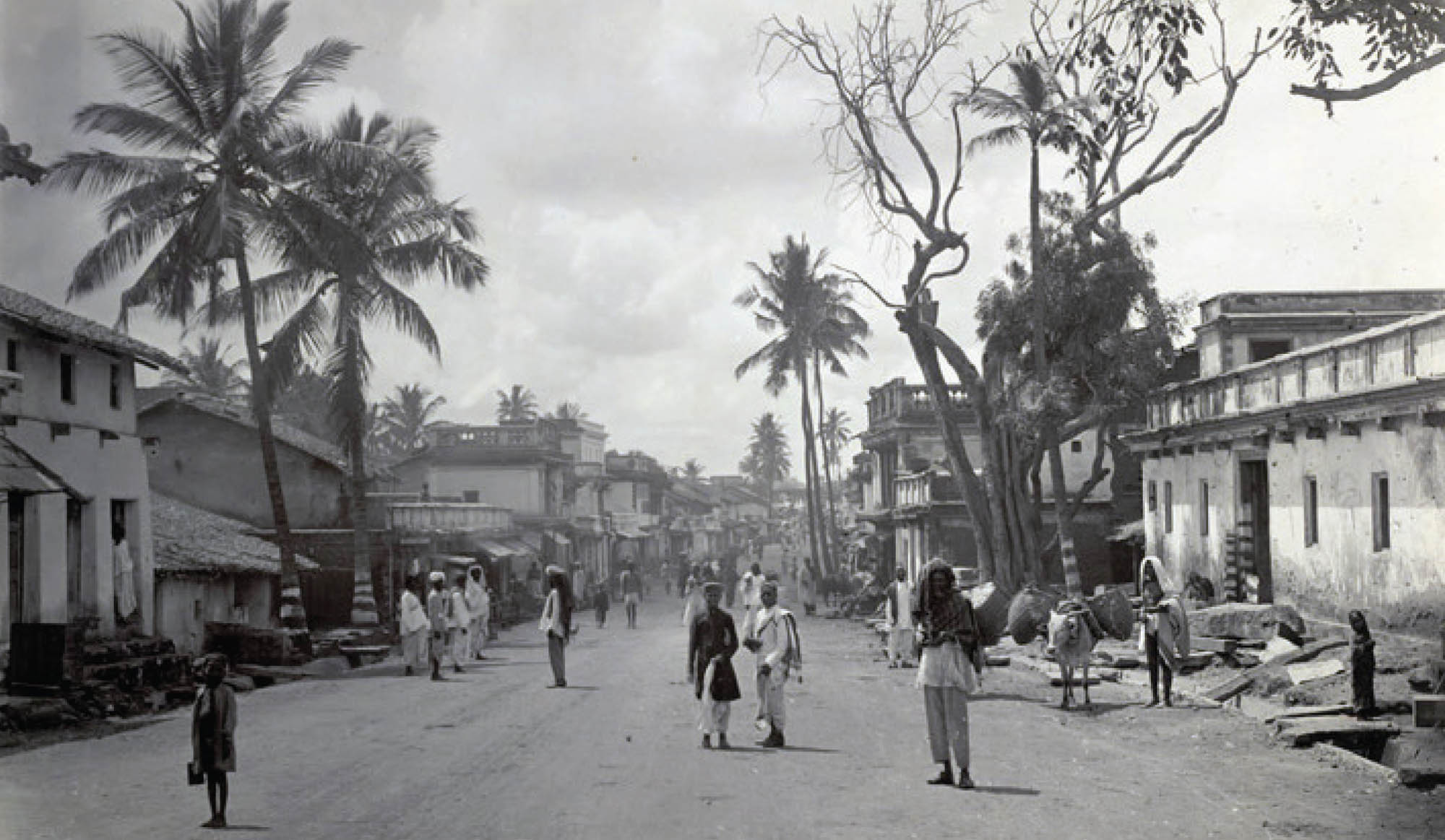 Black and white photograph of a city street in 1890 with pedestrians, trees, and shops.