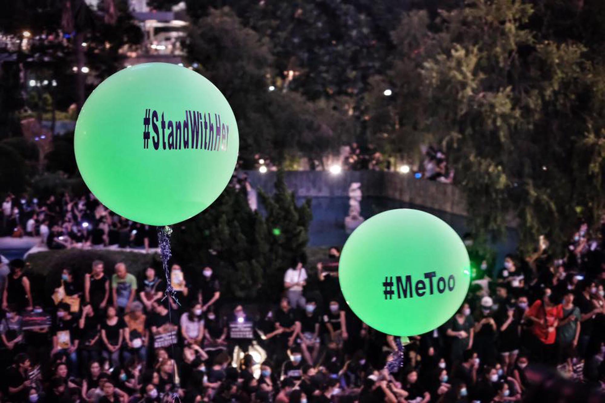 Two glowing green balloons, one labeled '#StandWithHer’ and the other '#MeToo', float above a crowd of people gathered at night in what appears to be a protest or rally.