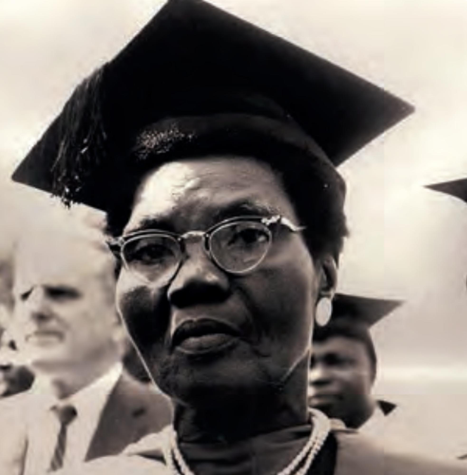 A close-up photo of Funmilayo Ransome-Kuti, wearing a graduation cap and gown, with a pearl necklace visible. She is looking directly at the camera with a serious expression, and other blurred figures are in the background.