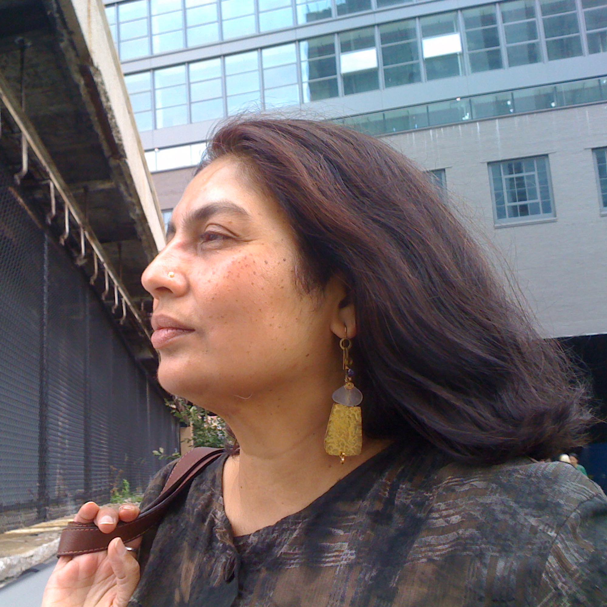 A side profile of Chandra Talpade Mohanty, she has dark hair and a nose ring and is looking intently to the left with a city building in the background.