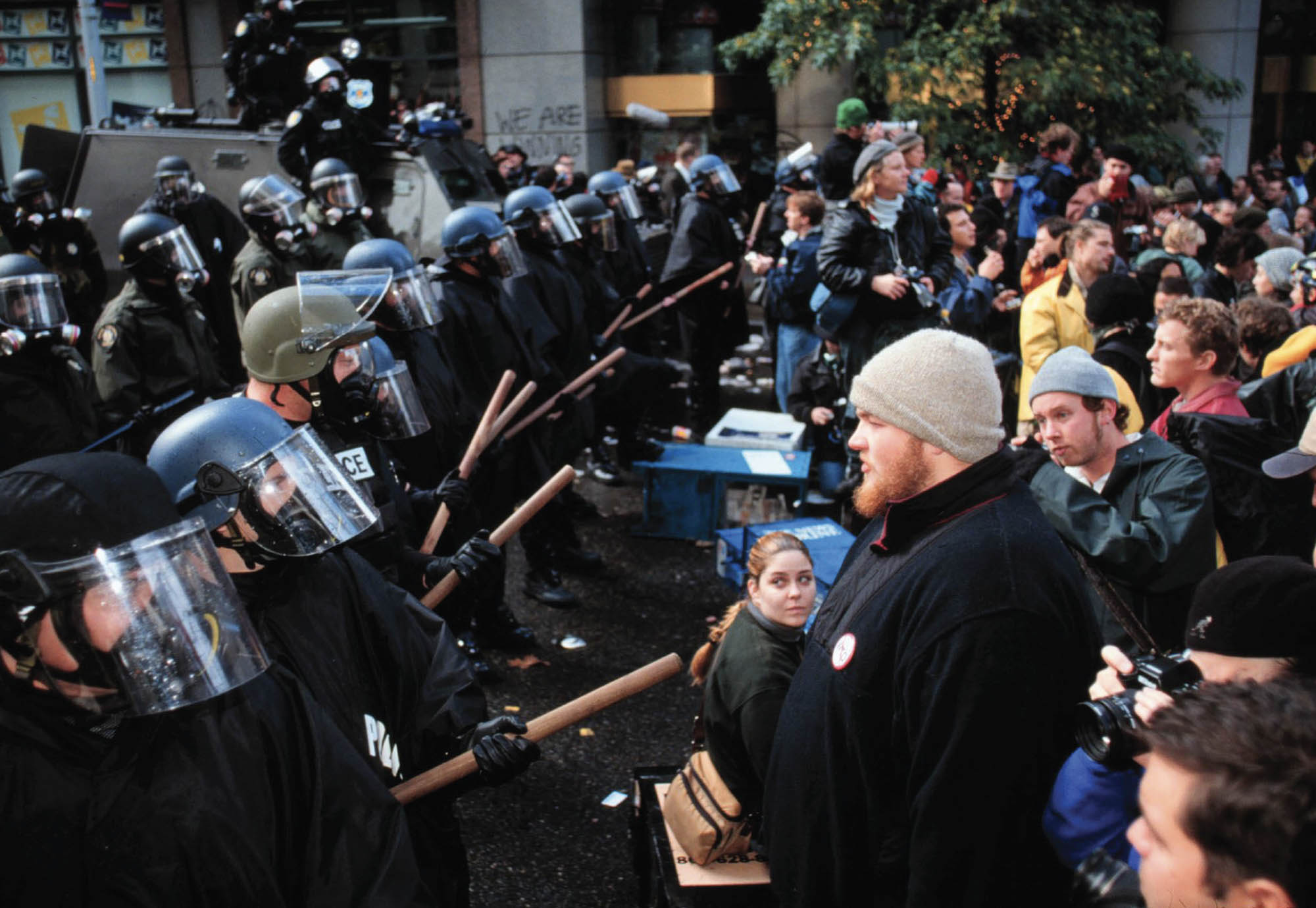 A photograph of a protest scene. A line of cops in full riot gear, wearing shields and holding batons face a line of protestors.