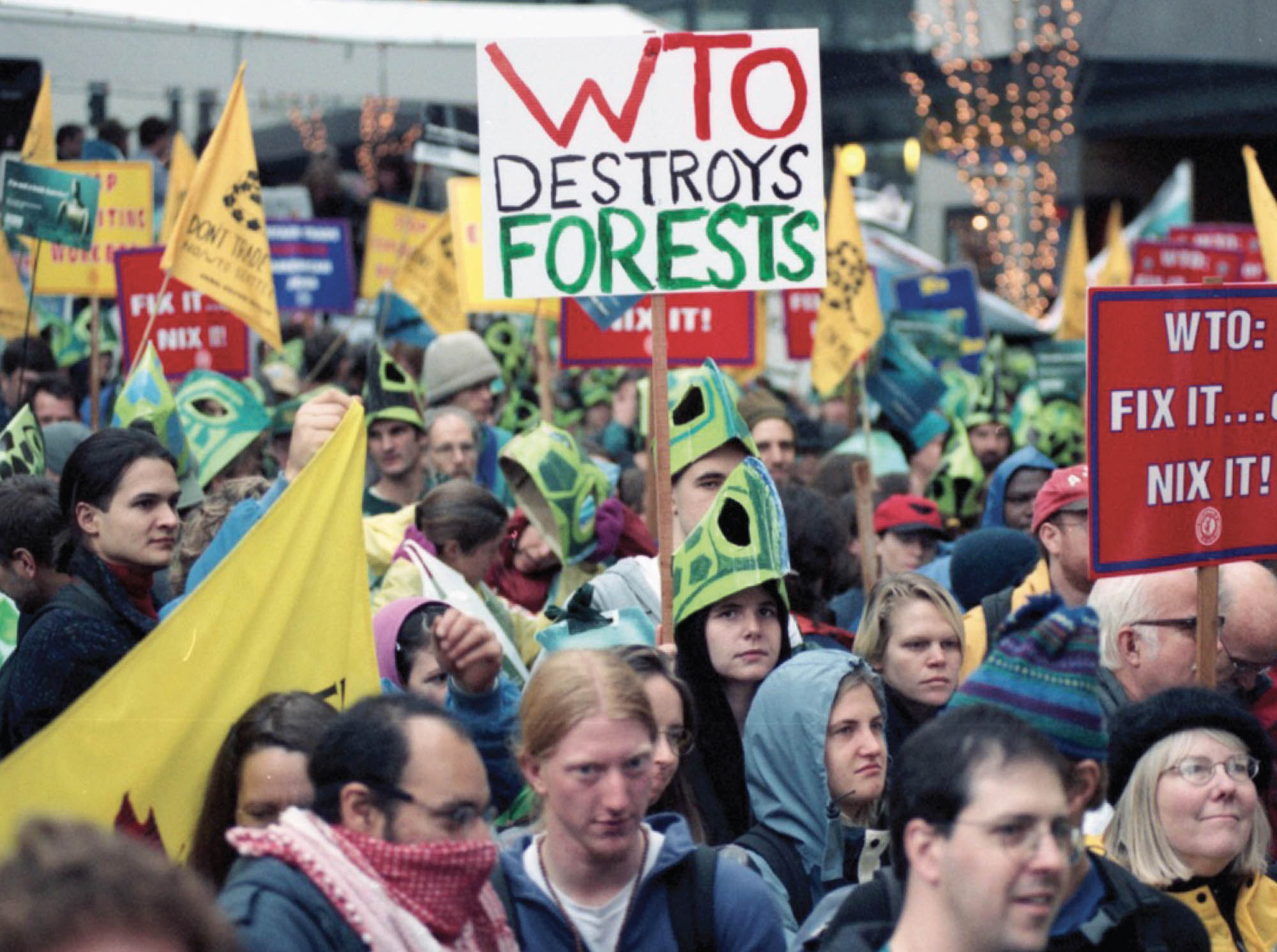 A crowd of protestors holding signs. One sign reads “WTO Destroys Forests”.