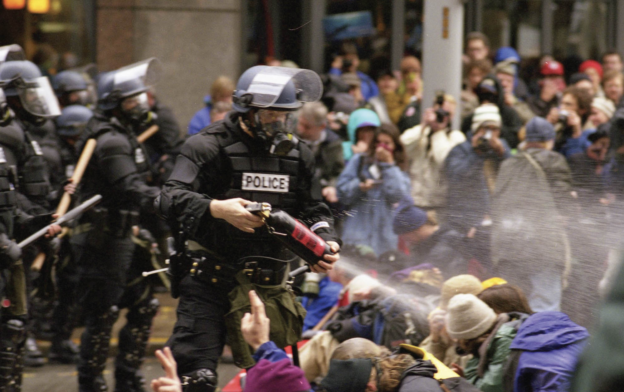 A photograph shows a police officer in full riot gear, flanked by several others, directly pepper-spraying several kneeling, peaceful protestors. The protestors are covering their faces and two of them are holding up peace signs with their hands despite being attacked.