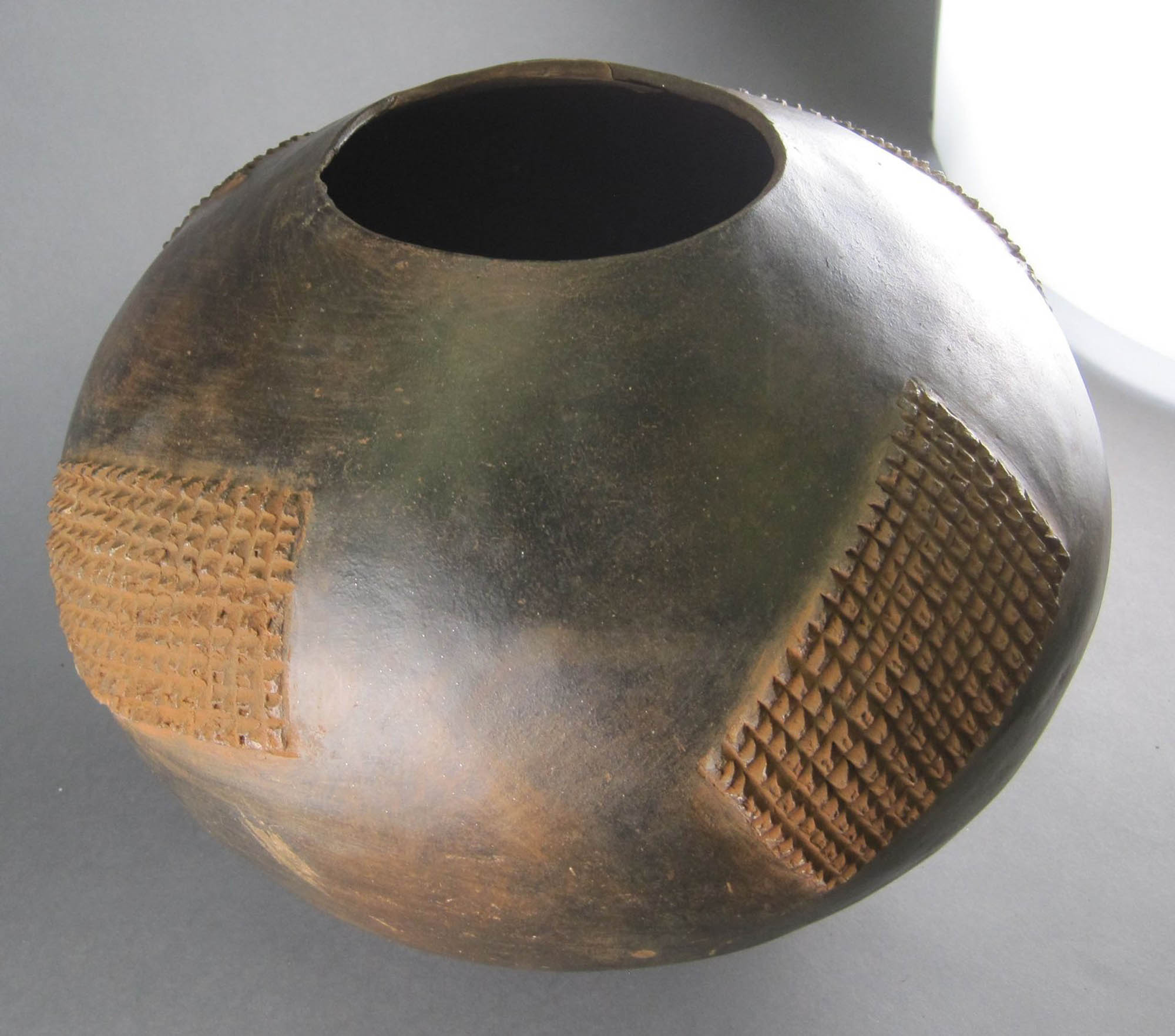 A large, round clay pot. There is a round opening on the top with decorative, textured rectangular shapes on the sides.