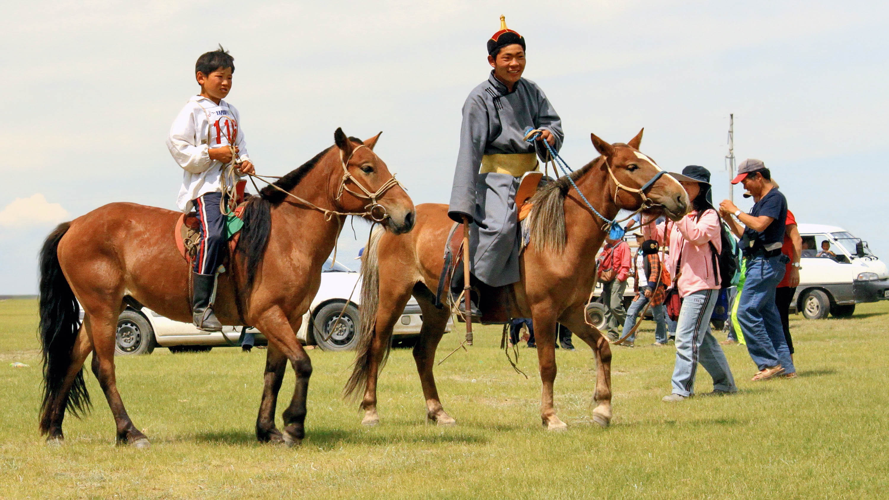 Two Mongolian riders, one adult and one child, on horseback in traditional dress, with people and vehicles in the background on an open grassy field.